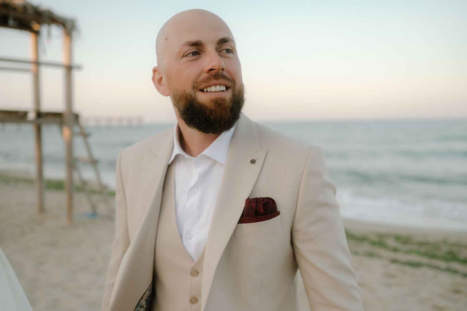 A beige suited figure stands smiling on a sunny beach with wooden posts and ocean waves in the background.