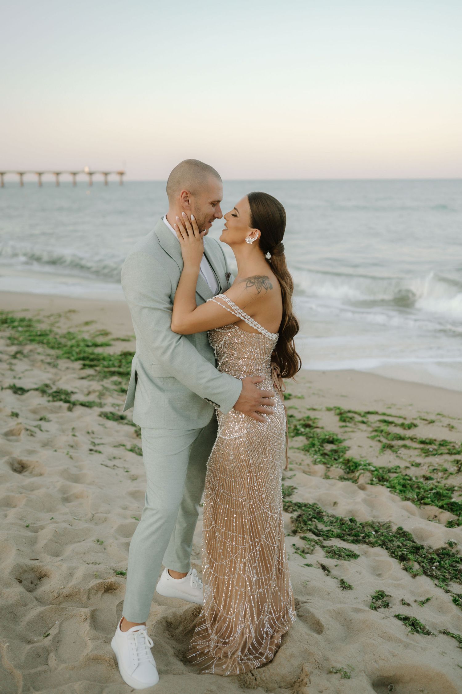 A couple embraces during their beachside photo session, with waves and a pier visible in the background.