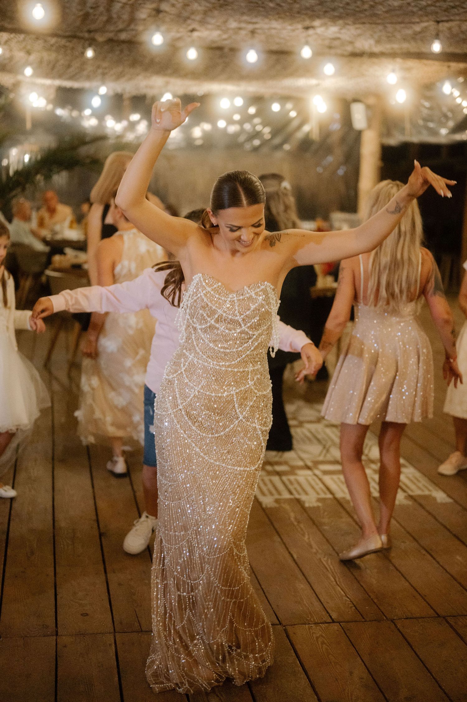 Wedding guests dance joyfully on a rustic wooden deck under glowing string lights at an evening reception.