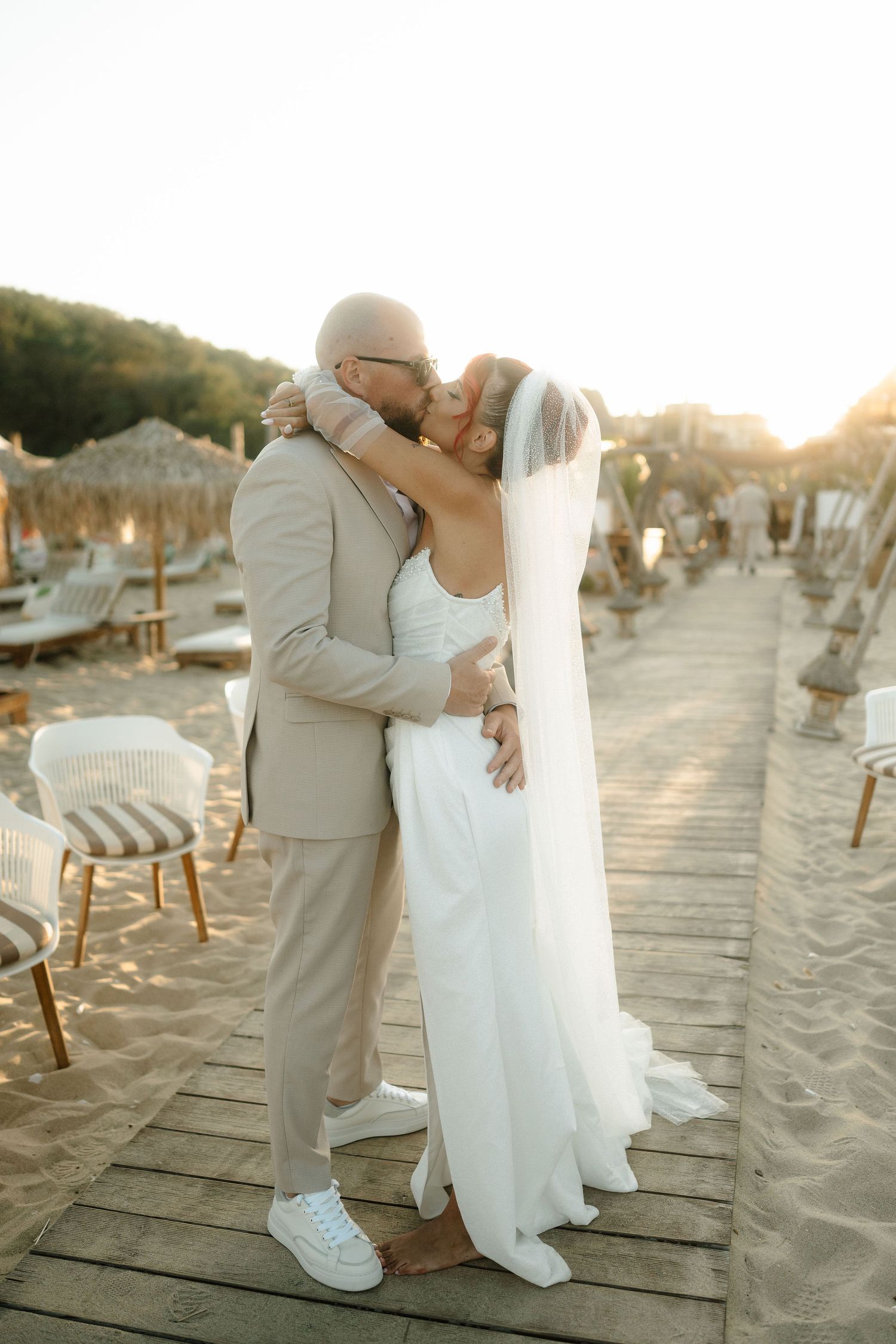 A romantic beach wedding couple embraces on a wooden boardwalk at sunset with sandy shores in the background.