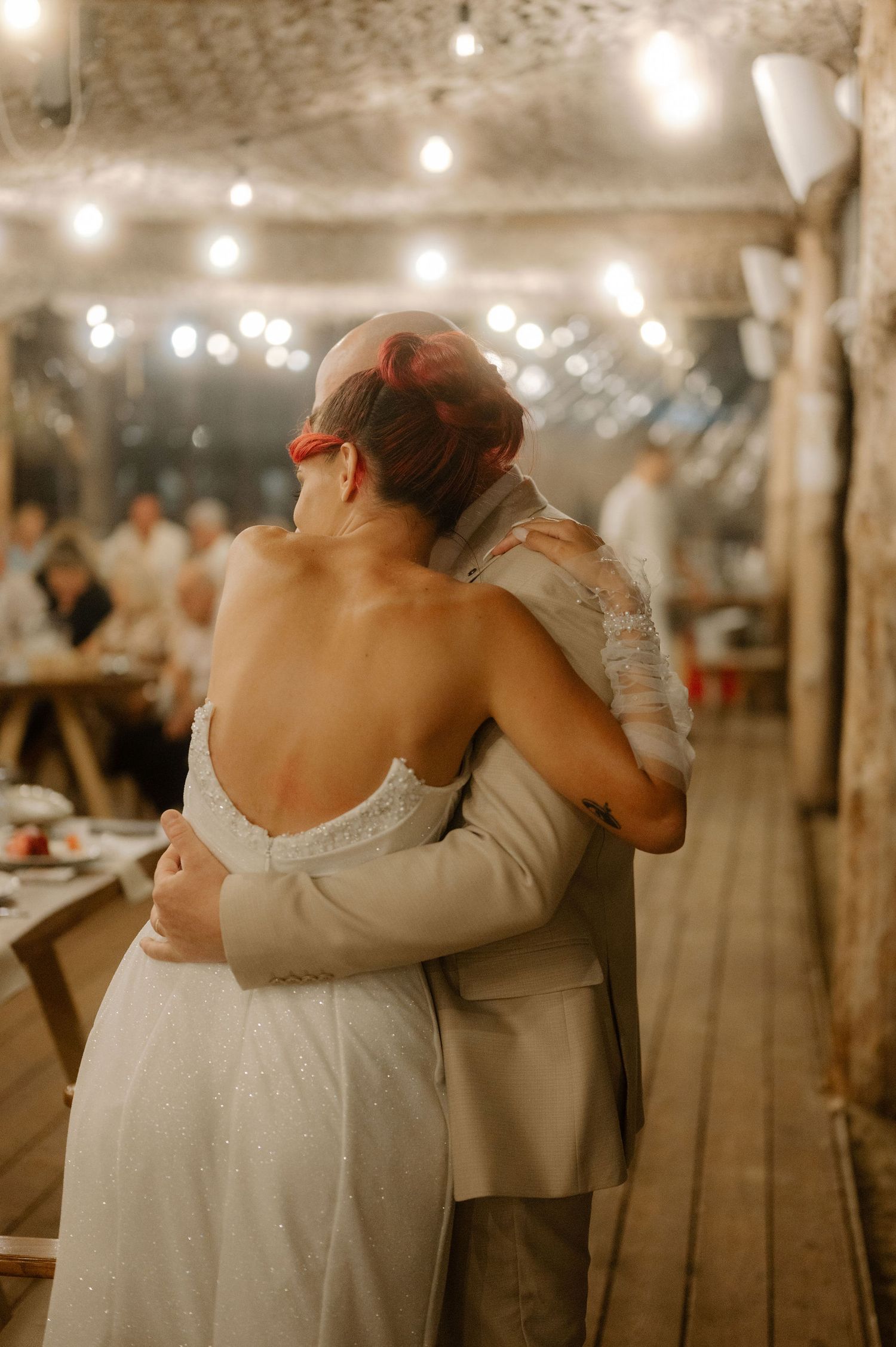 A couple embraces during a wedding reception under warm string lights in a rustic indoor venue.