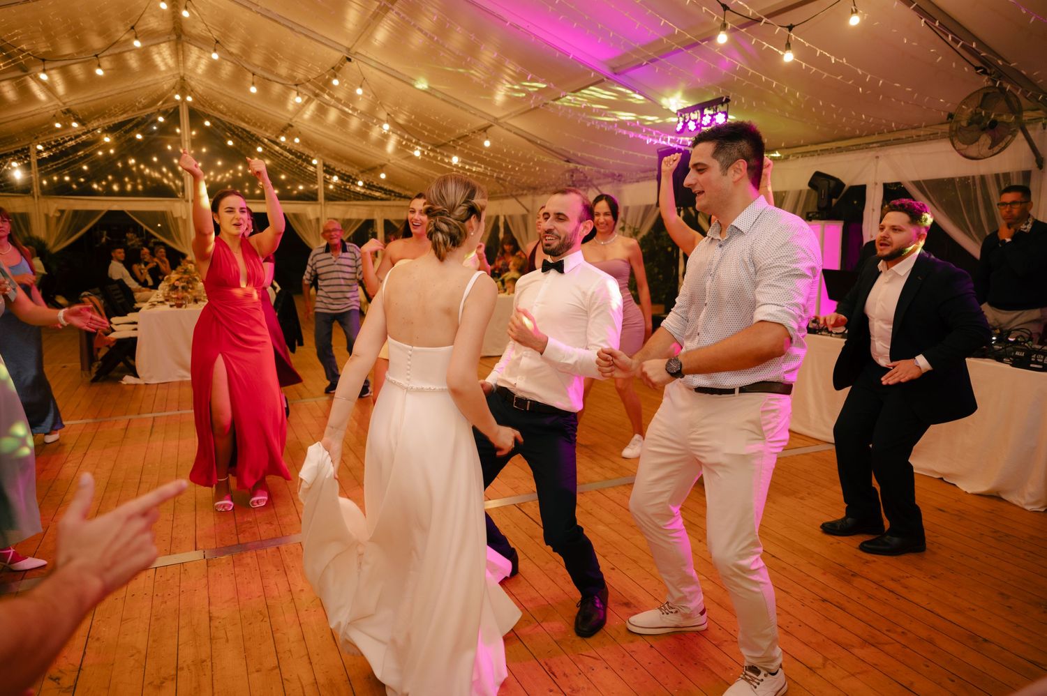 Couples dancing and celebrating at a wedding reception with pink lighting and string lights overhead.
