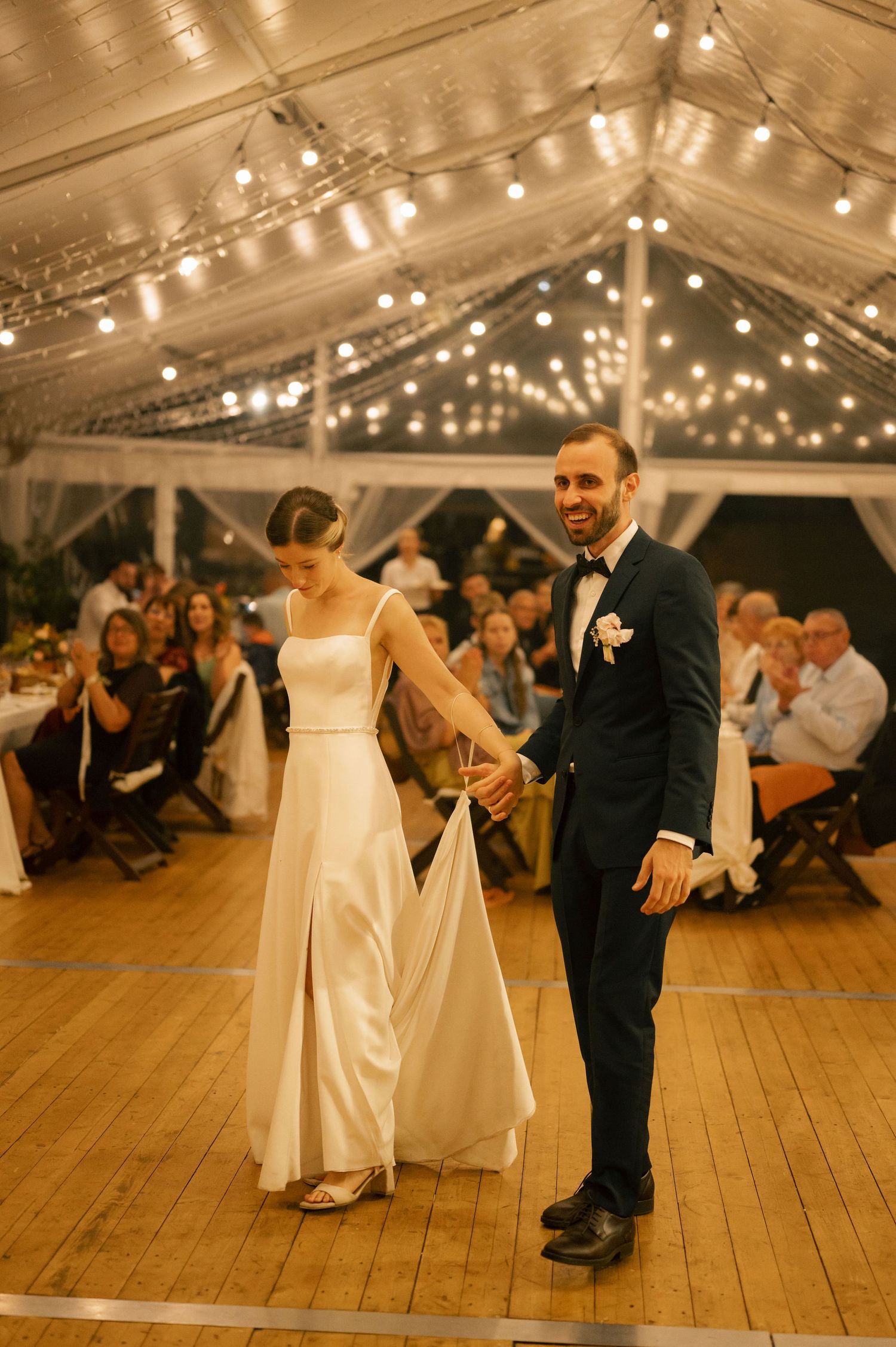 A couple shares a playful dance moment on a wooden floor under twinkling lights at a wedding reception.