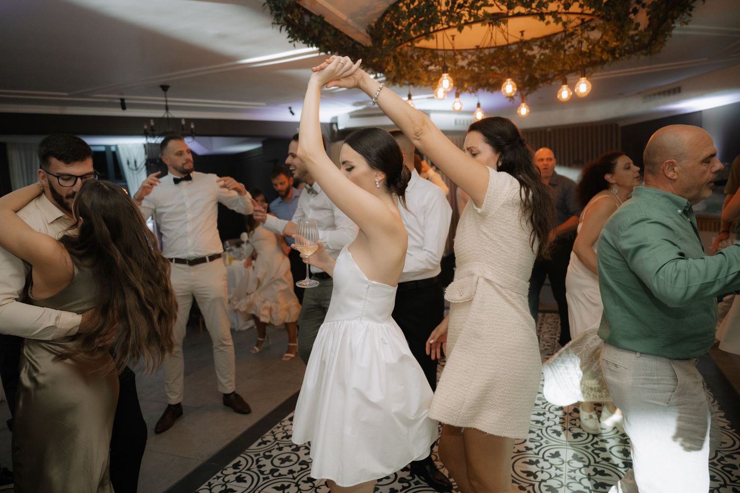 People dancing and celebrating at a wedding reception under decorative lighting on a patterned dance floor.