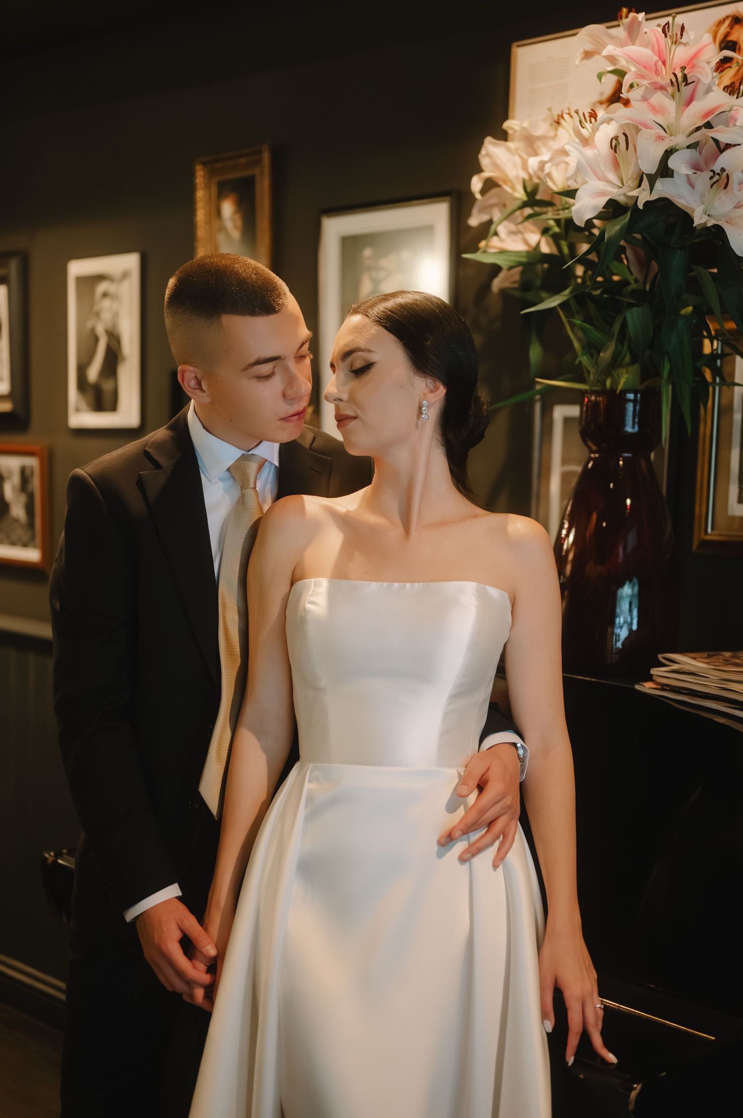 Romantic wedding couple shares intimate moments in an elegant dark room with framed artwork and floral arrangements.