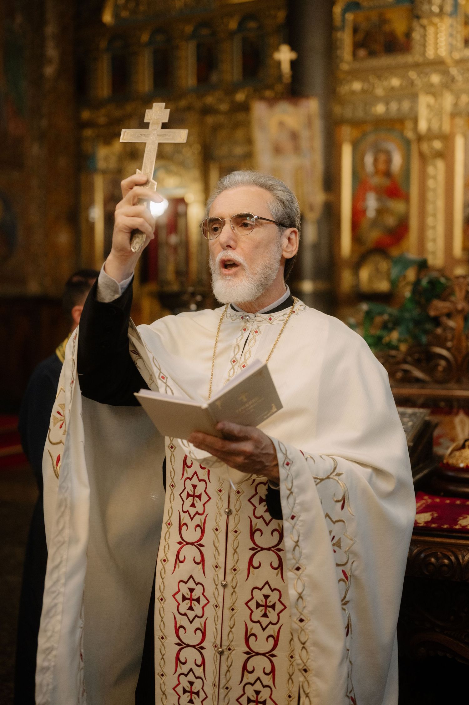 Orthodox priest in white embroidered vestments holds up a cross and book during religious ceremony in ornate church.