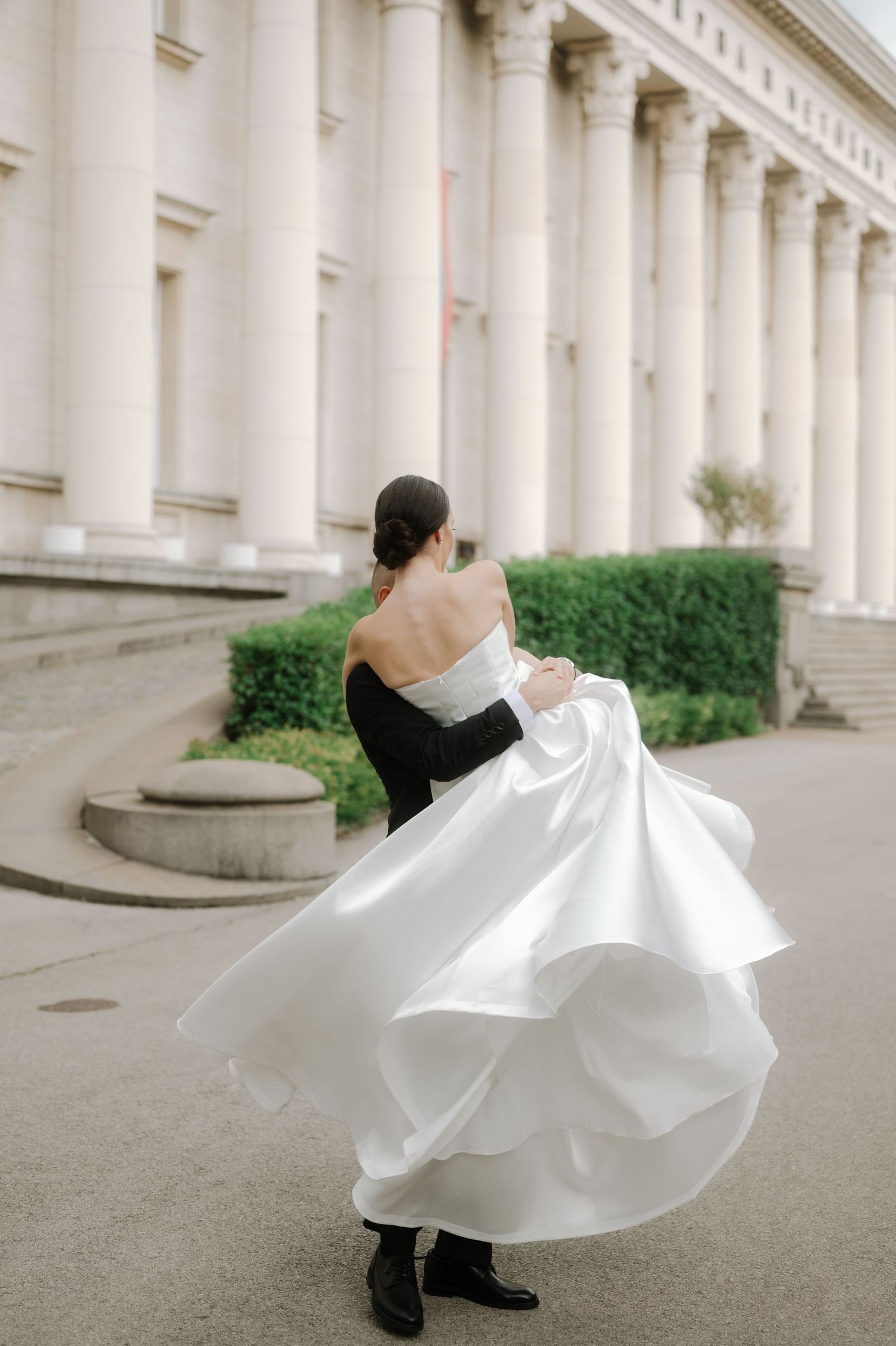 A person in a black and white formal dress twirls elegantly in front of a grand columned building.