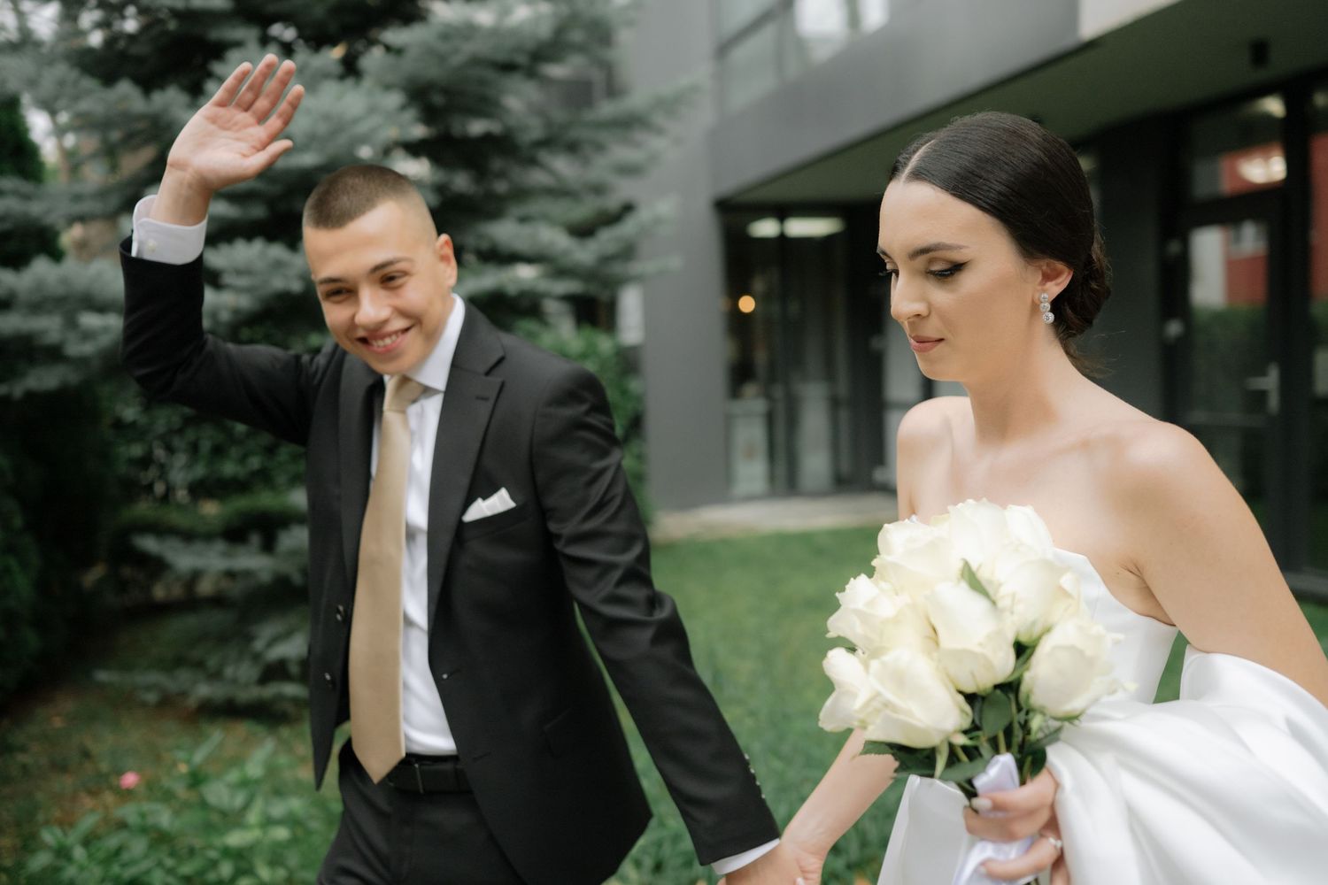 A couple in wedding attire walks outdoors, with the groom waving and bride holding white roses.