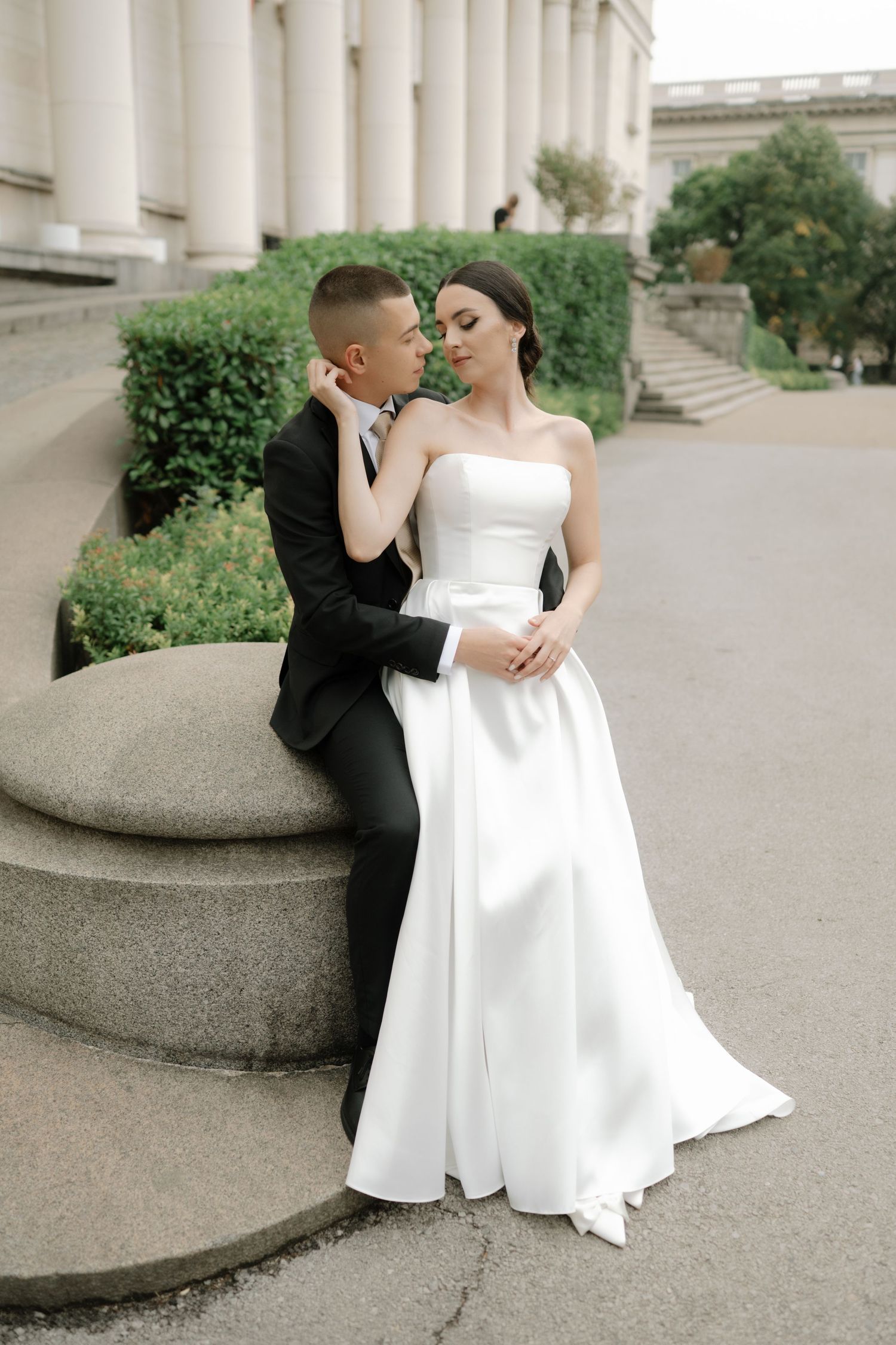 A couple in wedding attire share an intimate moment on stone steps outside a classical building with columns.