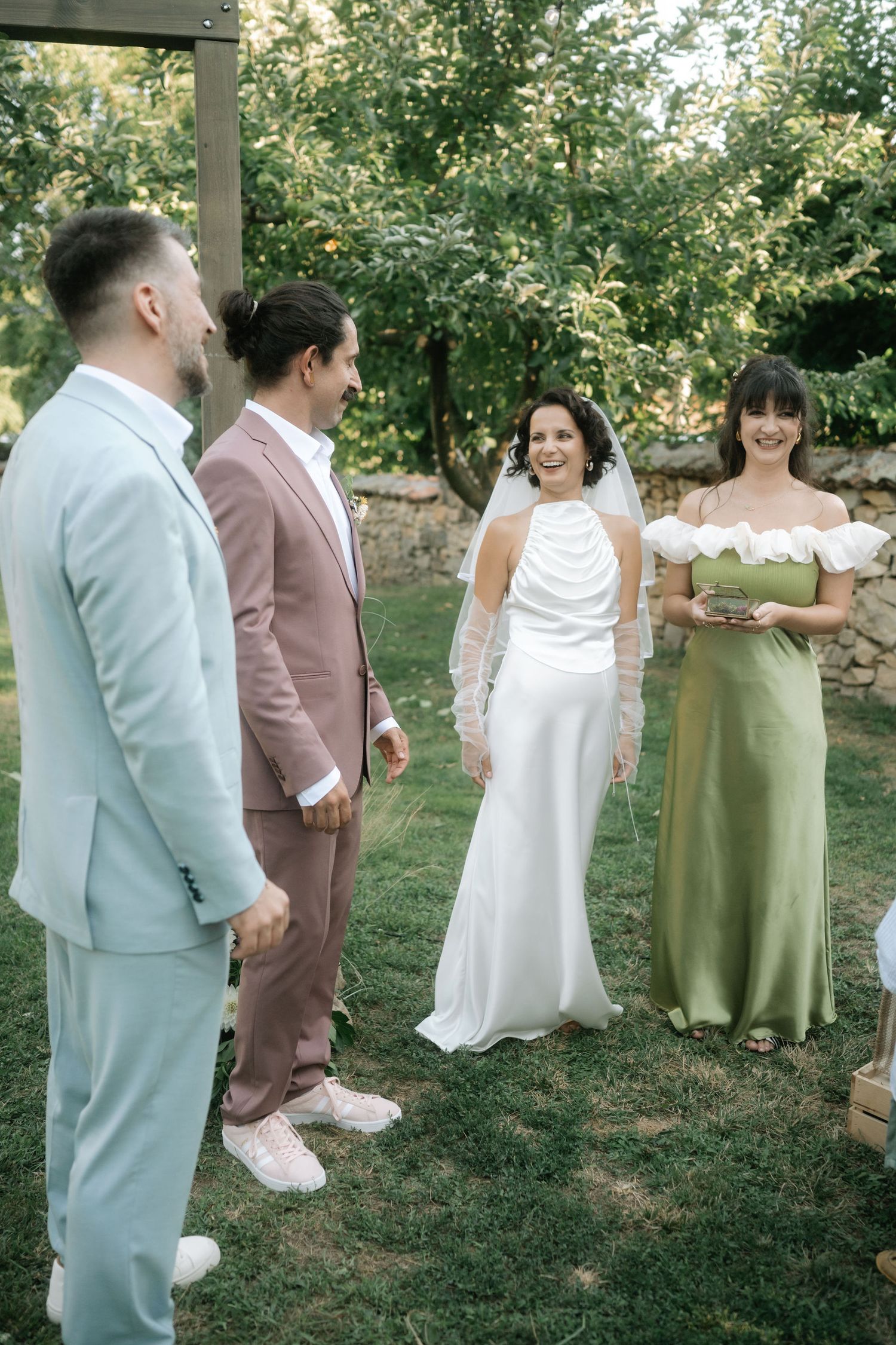 Wedding party members in coordinating pastel suits and dresses stand together on a lawn during an outdoor celebration.