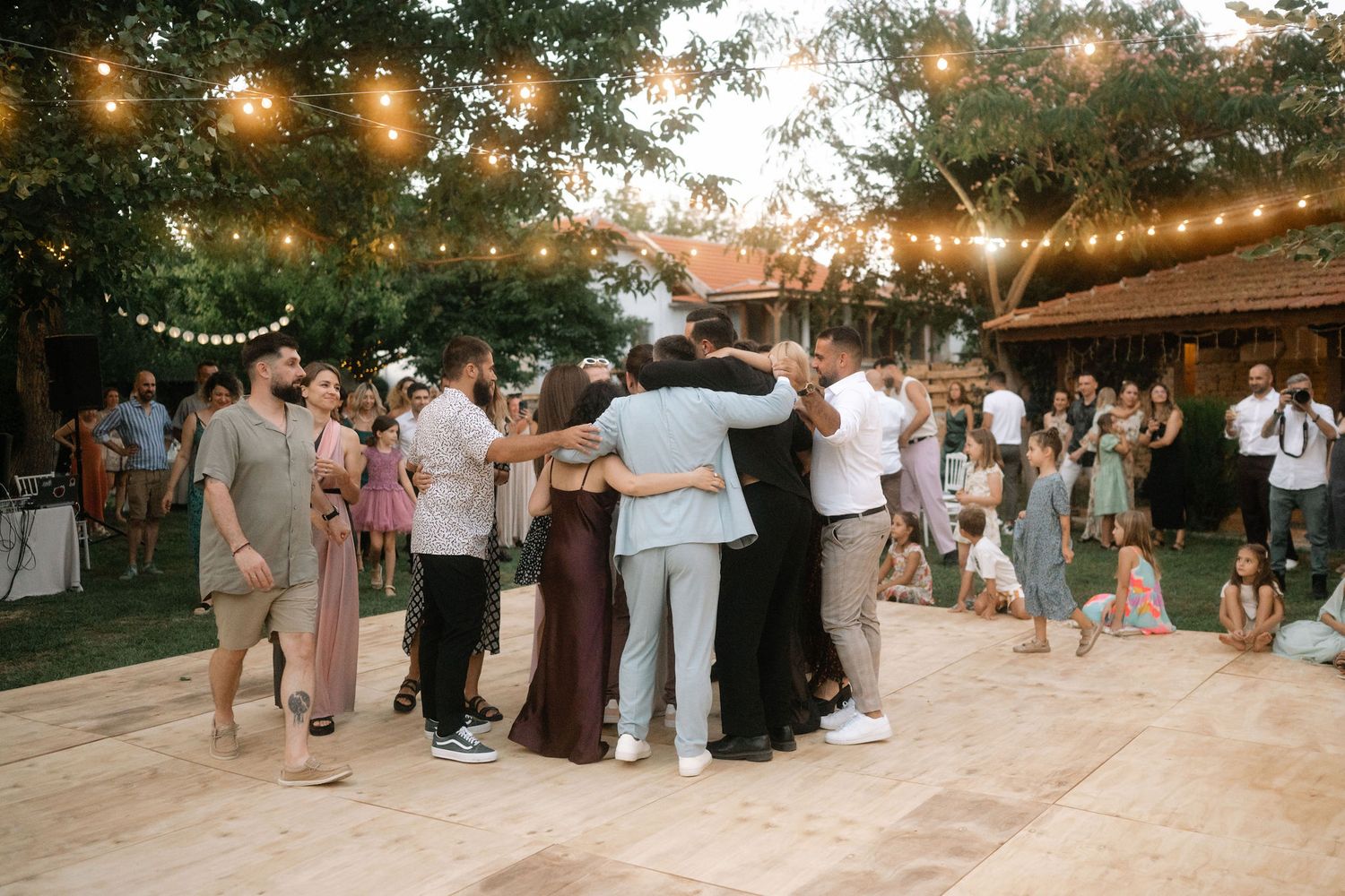 Guests gather outdoors at a rustic wedding reception venue with string lights and natural decor at dusk.