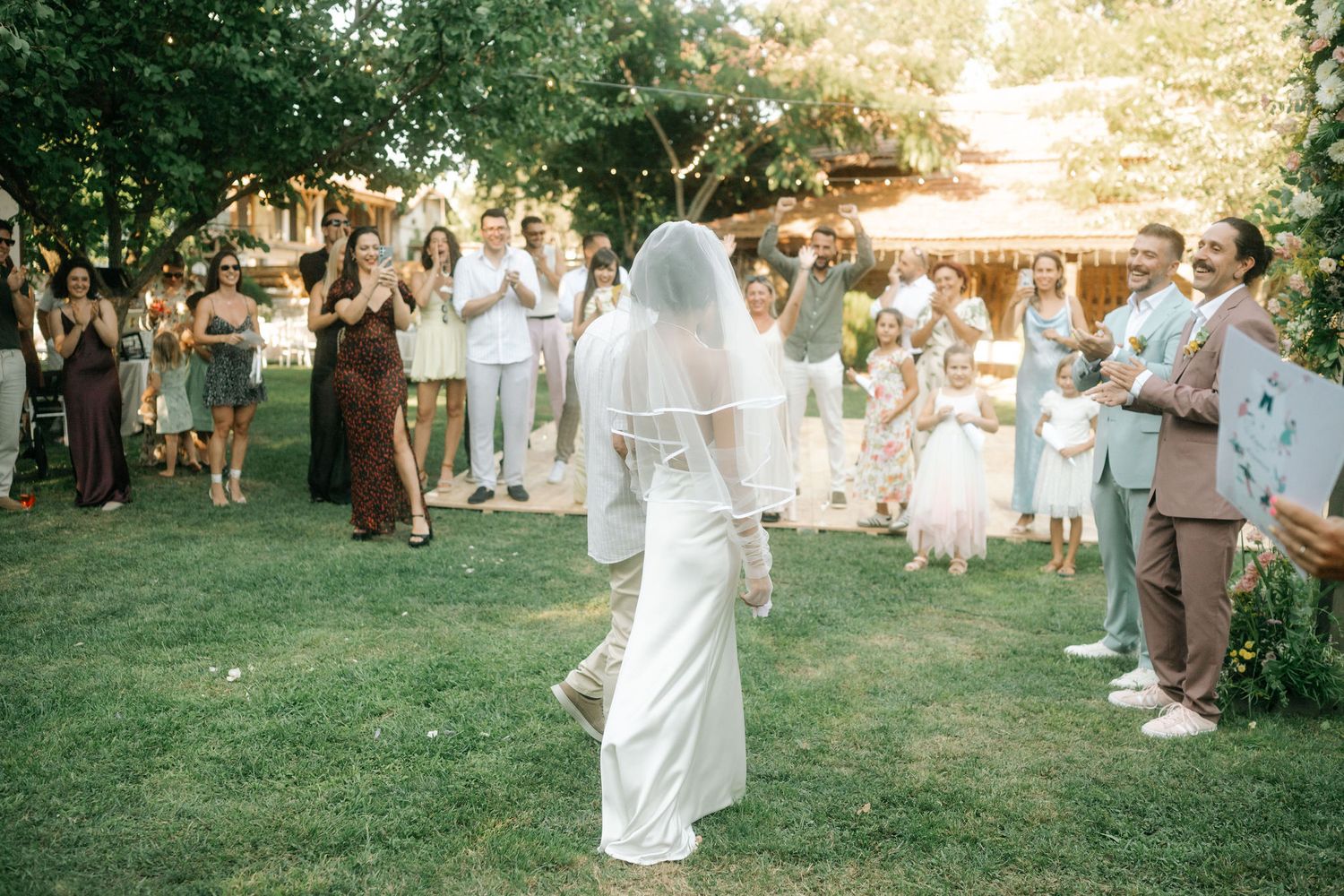 Outdoor wedding party dancing in a garden setting with golden sunlight filtering through trees.