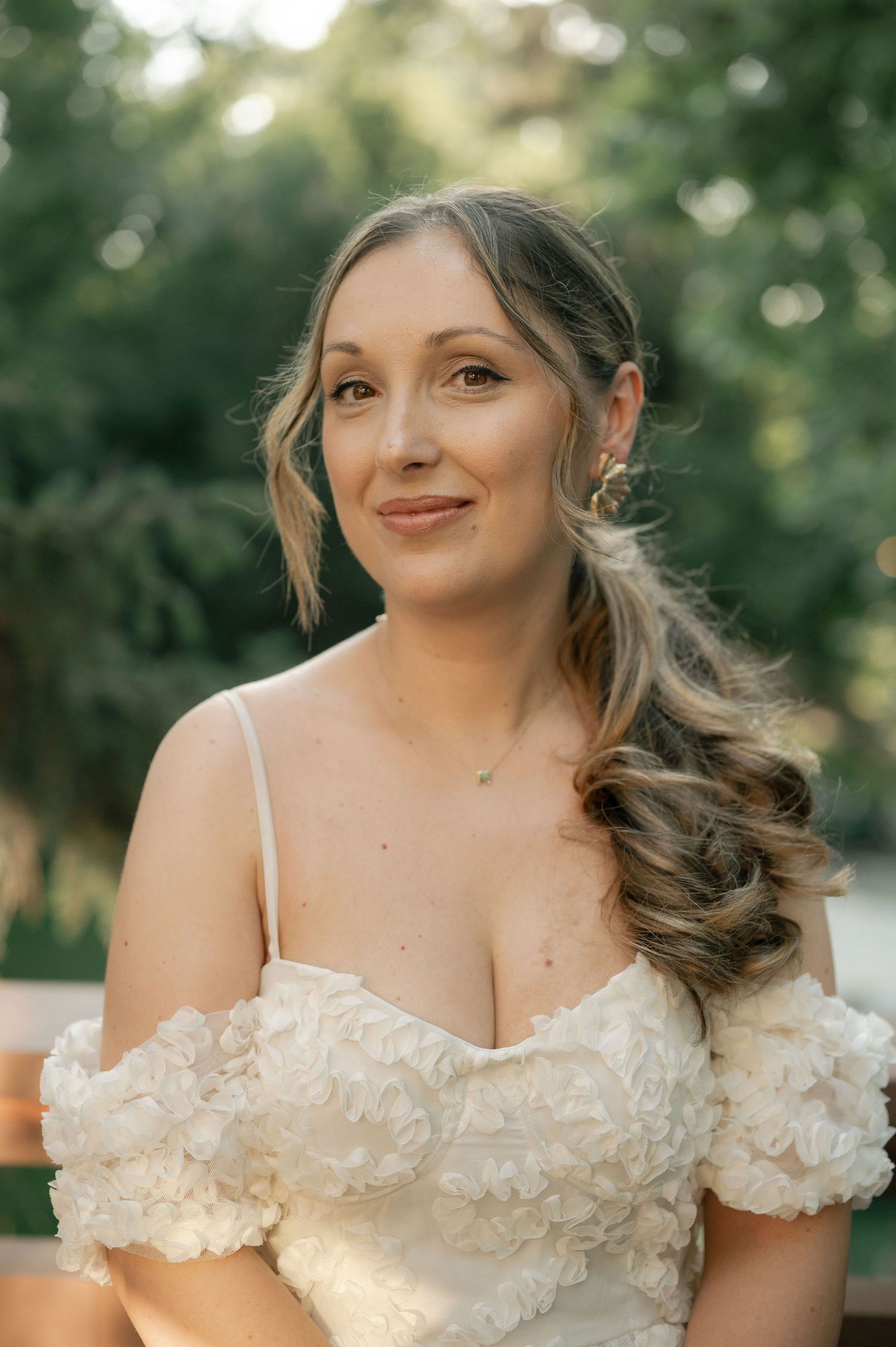 A bride in an off-shoulder lace wedding dress poses outdoors with soft bokeh lighting and styled wavy hair.