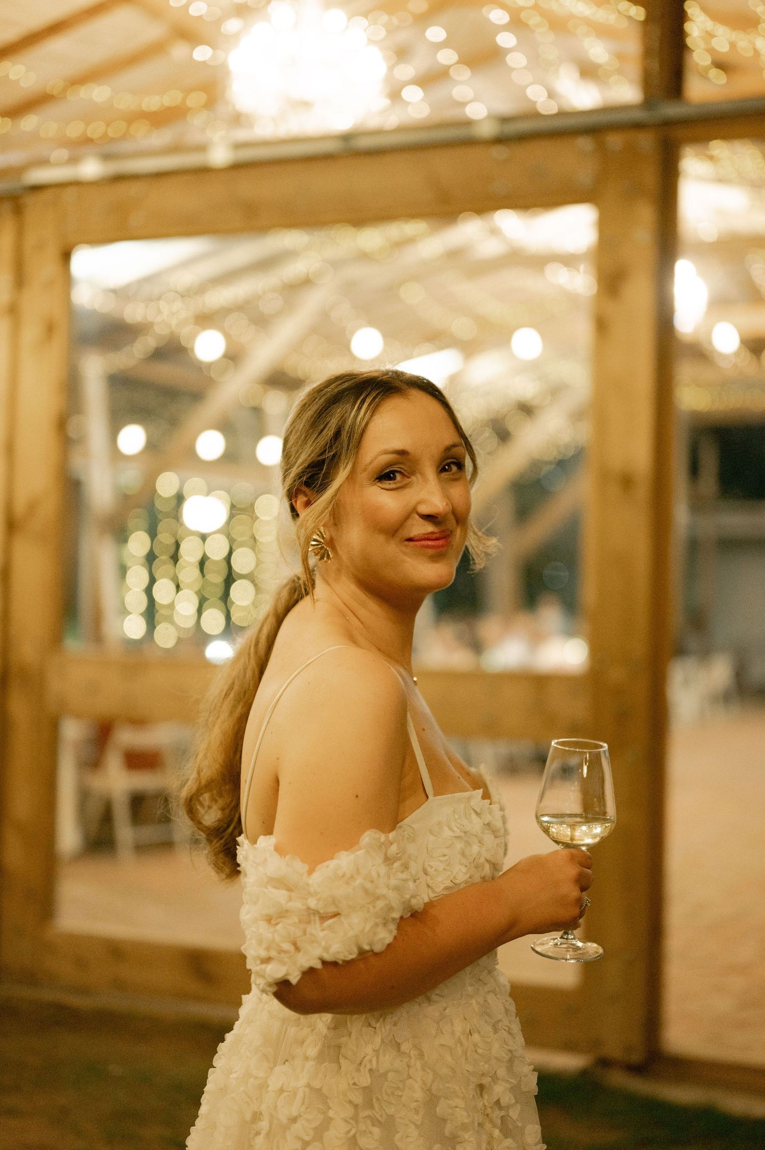 A bride in a white lace wedding dress holds a wine glass while smiling in a warmly lit reception venue.