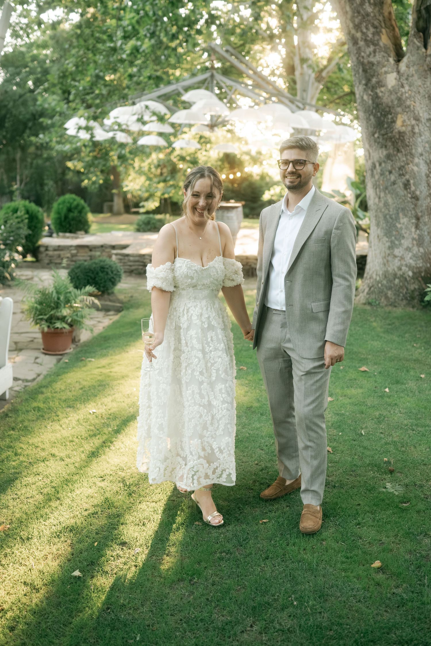 A couple in wedding attire stands on a sunlit garden lawn with trees and potted plants in the background.