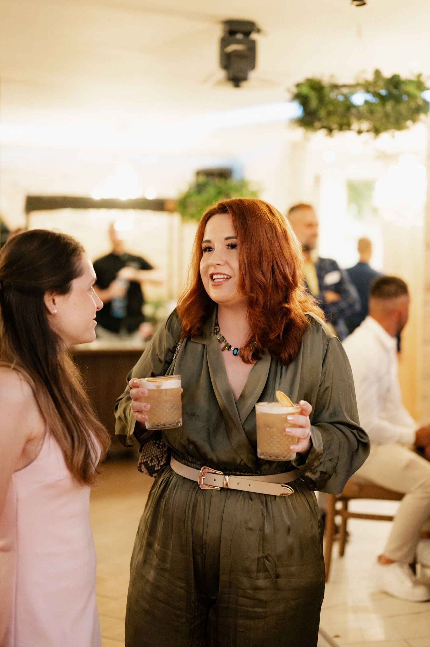 Two people in casual attire having an animated conversation while holding drinks at an indoor gathering.