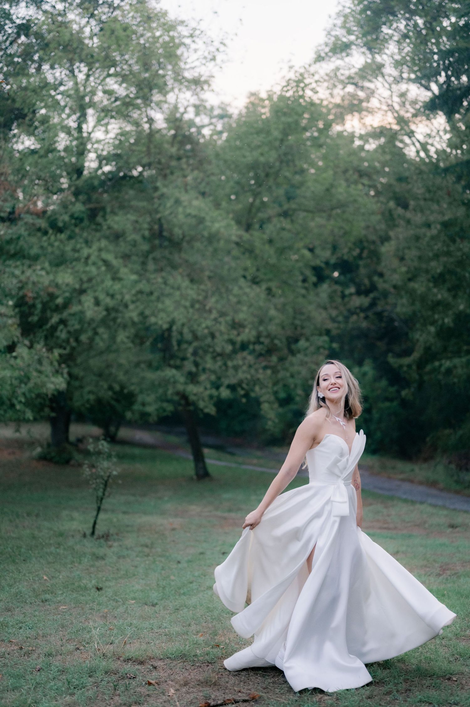 A bride spins joyfully in her elegant white gown, making the dress billow out against a backdrop of trees.