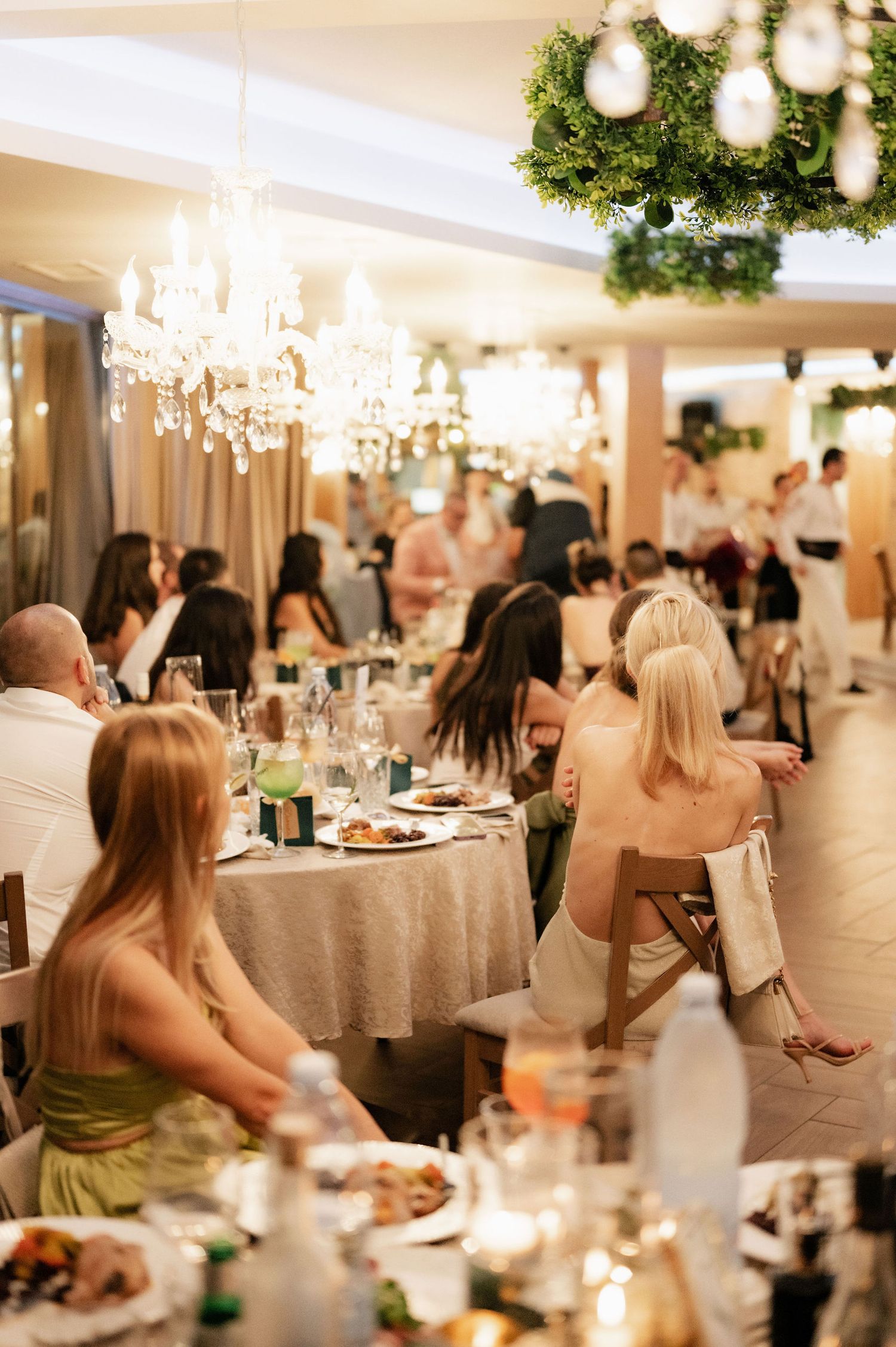 Wedding reception guests seated at elegant round tables with greenery decor and chandeliers in a bright ballroom setting.