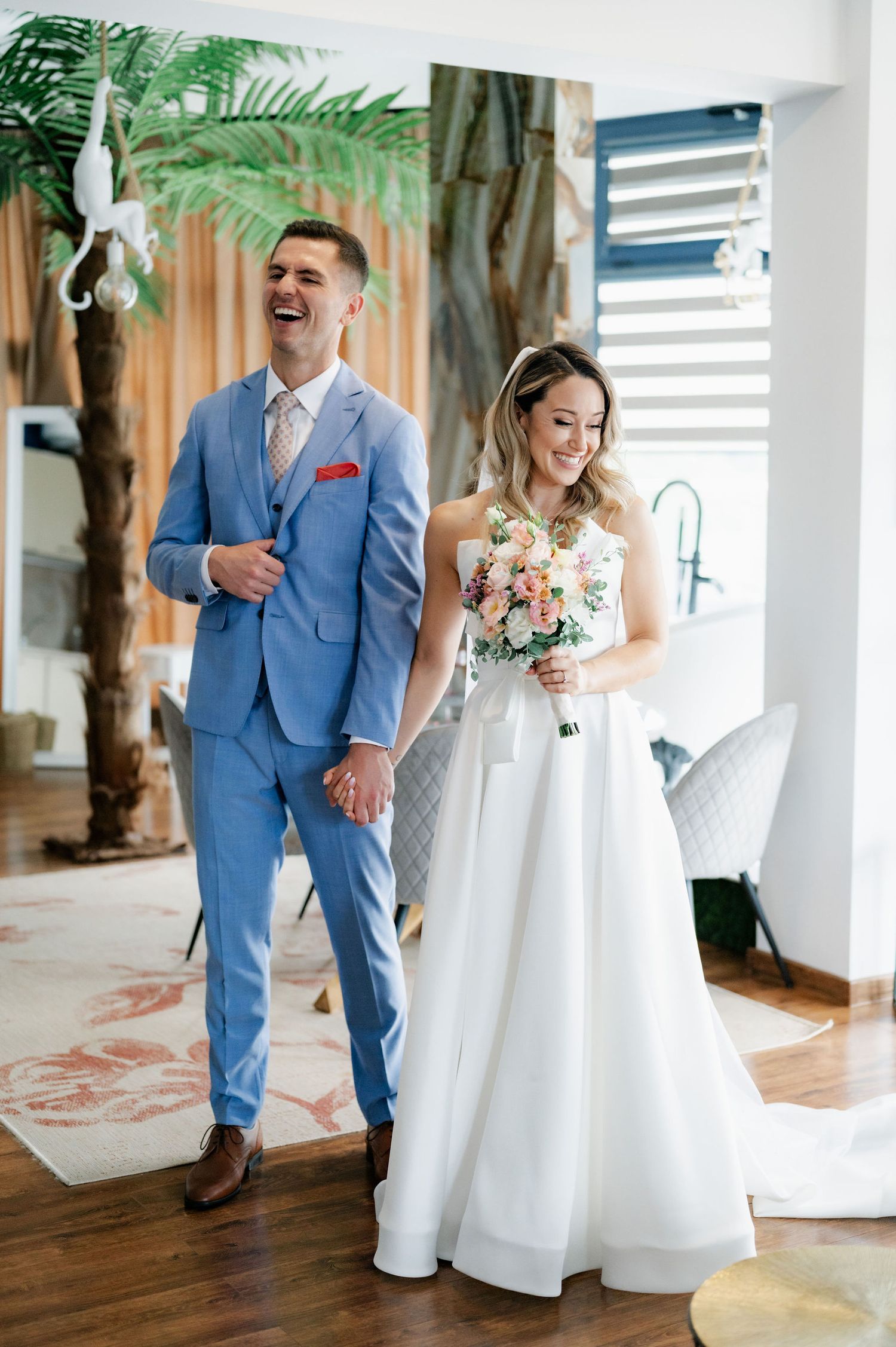 A couple in wedding attire share a joyful moment in a bright indoor venue with tropical plants and wooden floors.