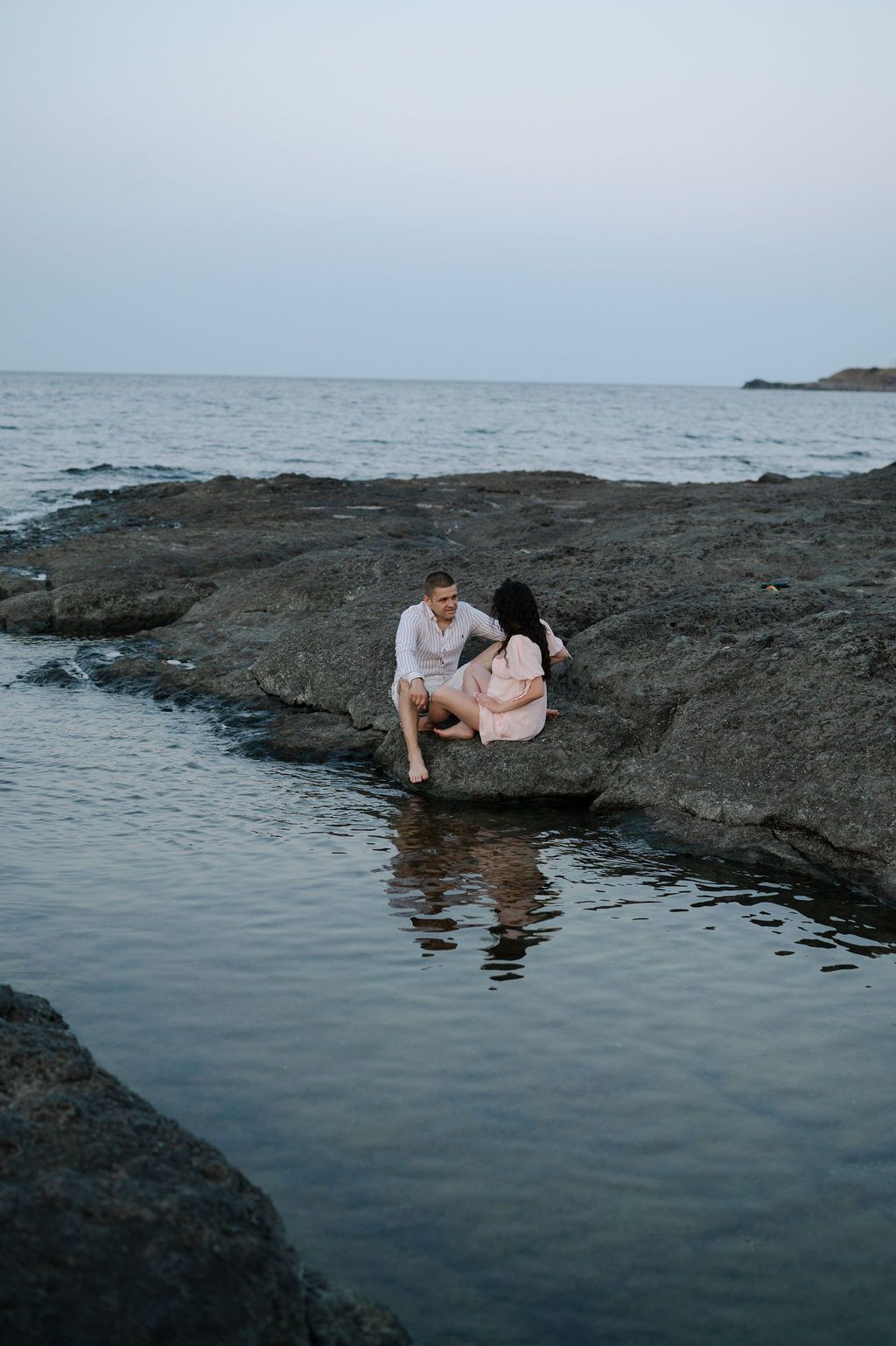 Person relaxing on dark volcanic rocks by calm ocean water at sunset, creating peaceful reflection in tidal pool.