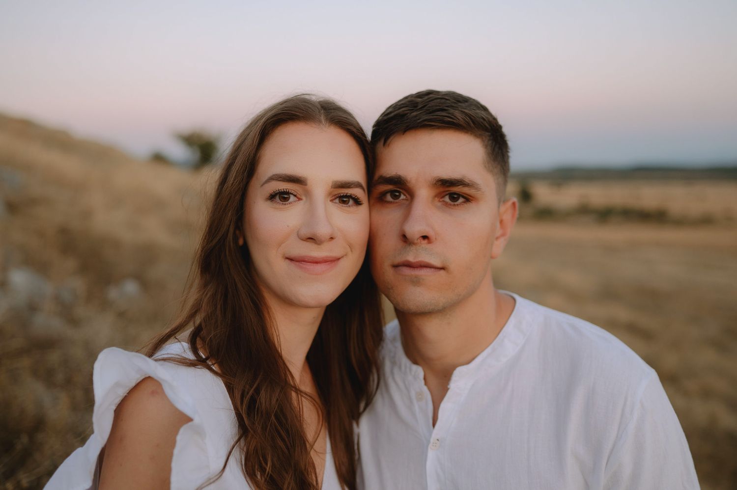 A couple embraces during a sunset photoshoot in a golden field with warm lighting and natural scenery.