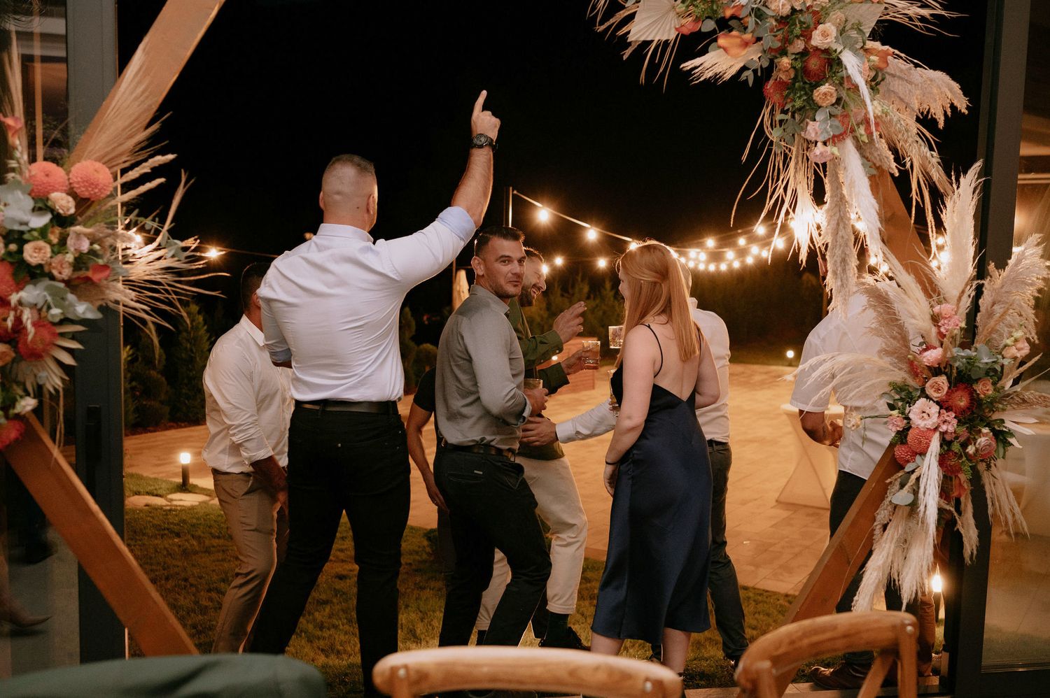 People dancing at a nighttime wedding reception under string lights and pampas grass decorations.