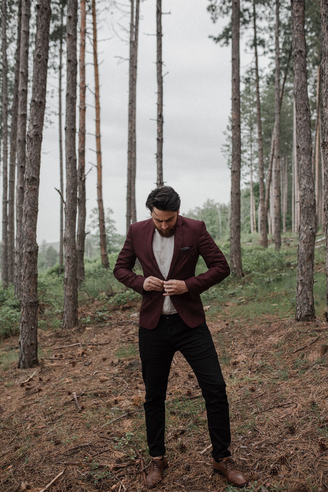 A person in a maroon suit jacket and dark pants stands on a forest slope among tall pine trees.