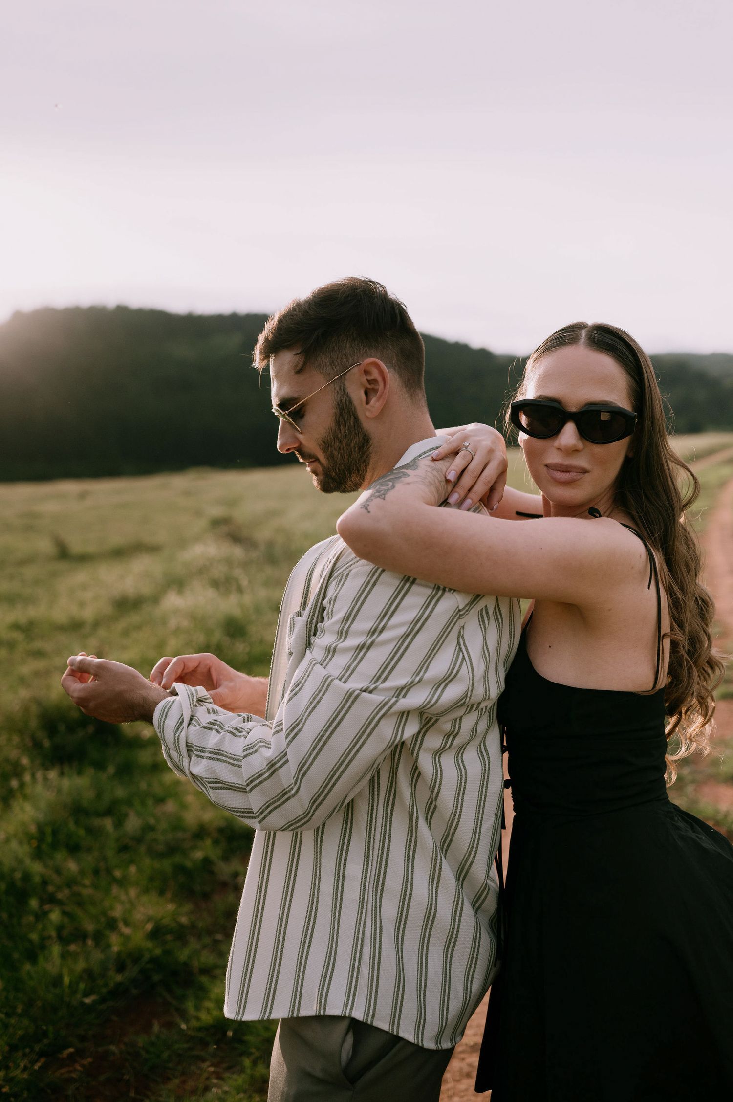 A couple embraces in a scenic mountain field during golden hour, with sunlight creating a warm glow behind them.