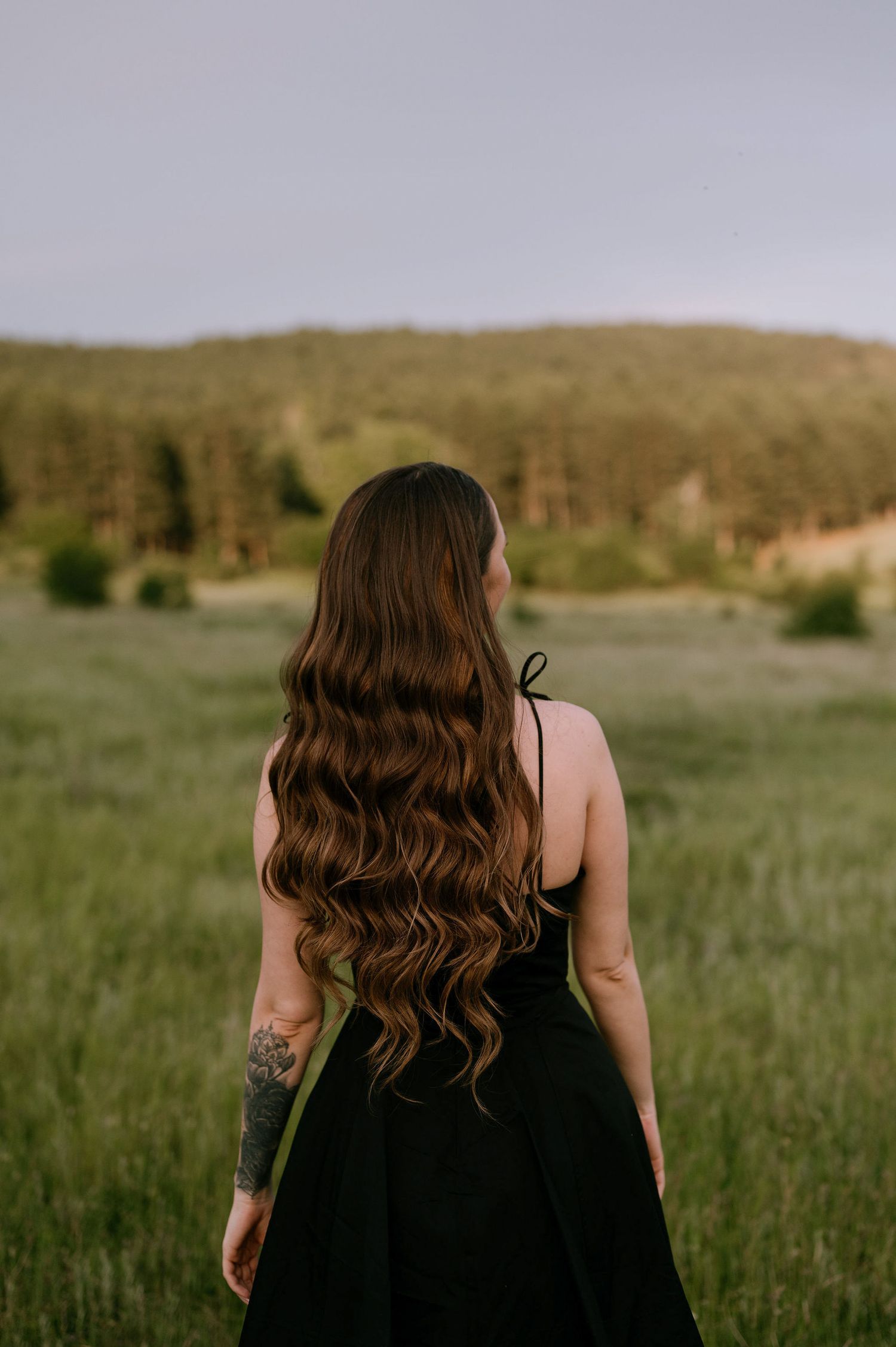 Person with long wavy brown hair stands in a grassy meadow with rolling hills in the background at sunset.