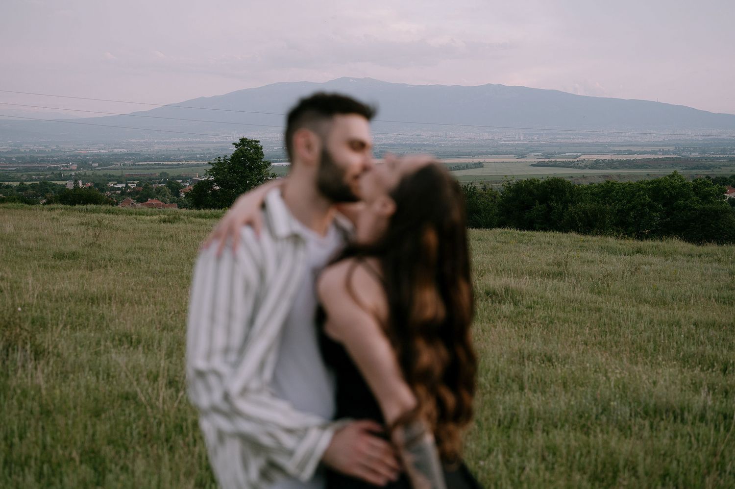 Romantic couple embraces in a serene meadow with rolling hills and mountains in the background at sunset.