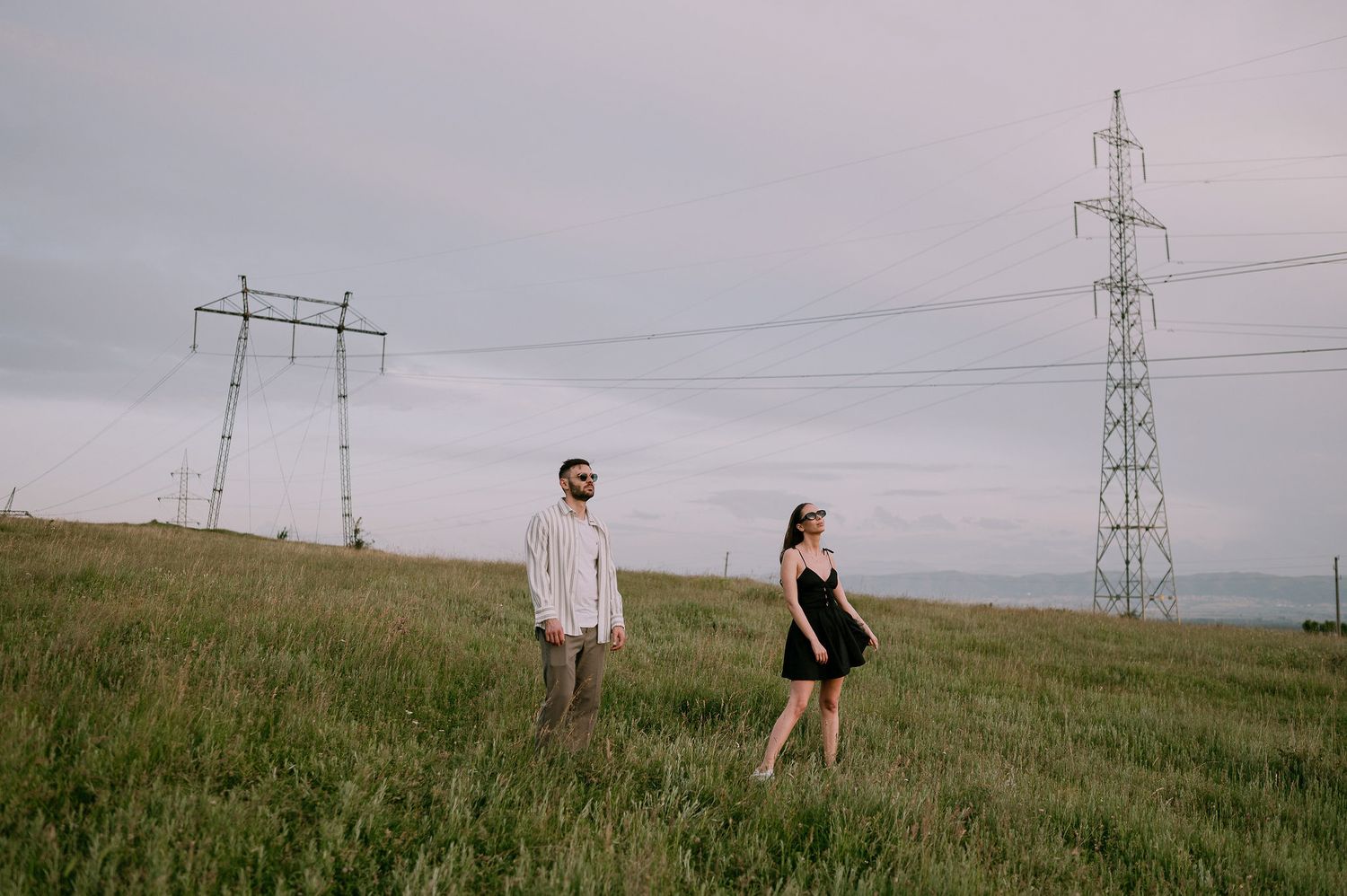 People walk through a grassy field with electrical transmission towers stretching into the distance on an overcast day.