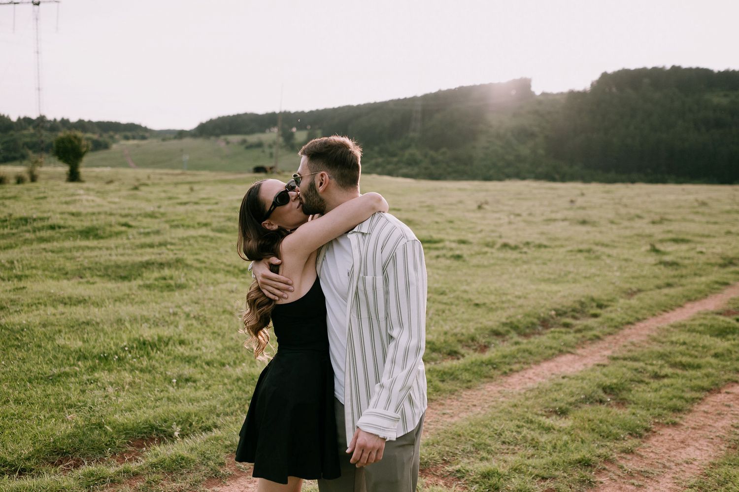 A couple shares a romantic embrace in a sunlit meadow with a dirt path and rolling hills in the background.
