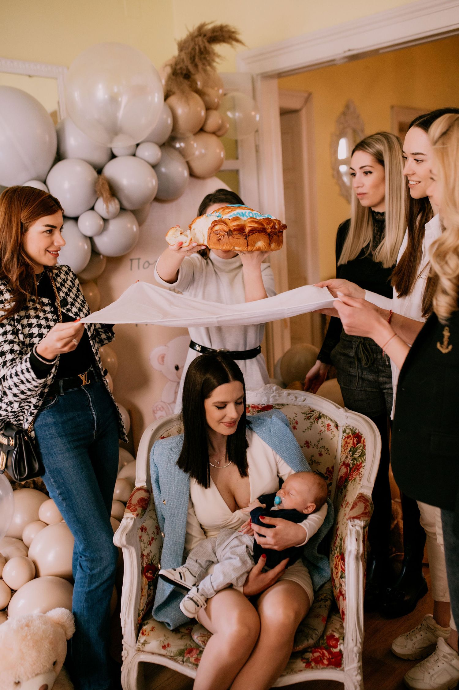 A group gathers in a room decorated with white balloons and soft decor for a baby shower celebration.