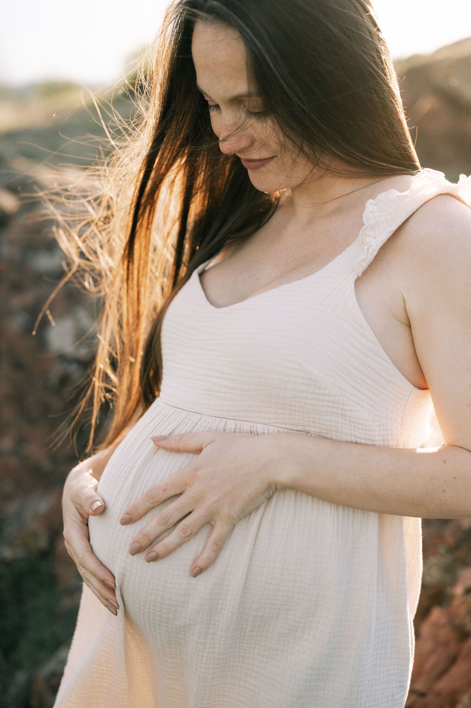 Expectant mother in white dress holds her baby bump while standing in warm sunset light with wind-blown hair.