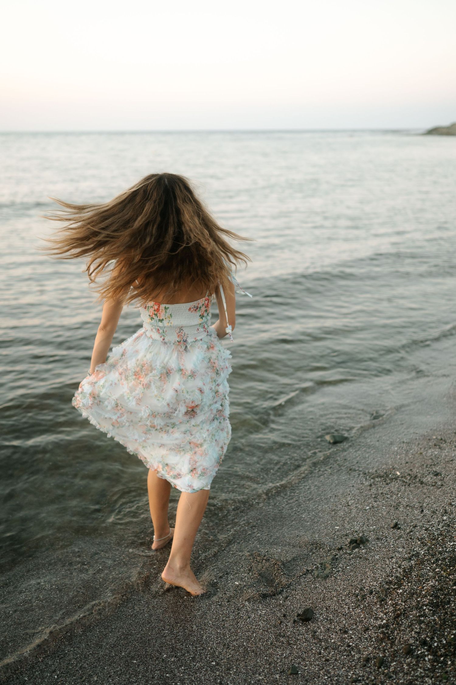 A person in a flowy floral dress dances along the shoreline at sunset, creating a dreamy coastal scene.