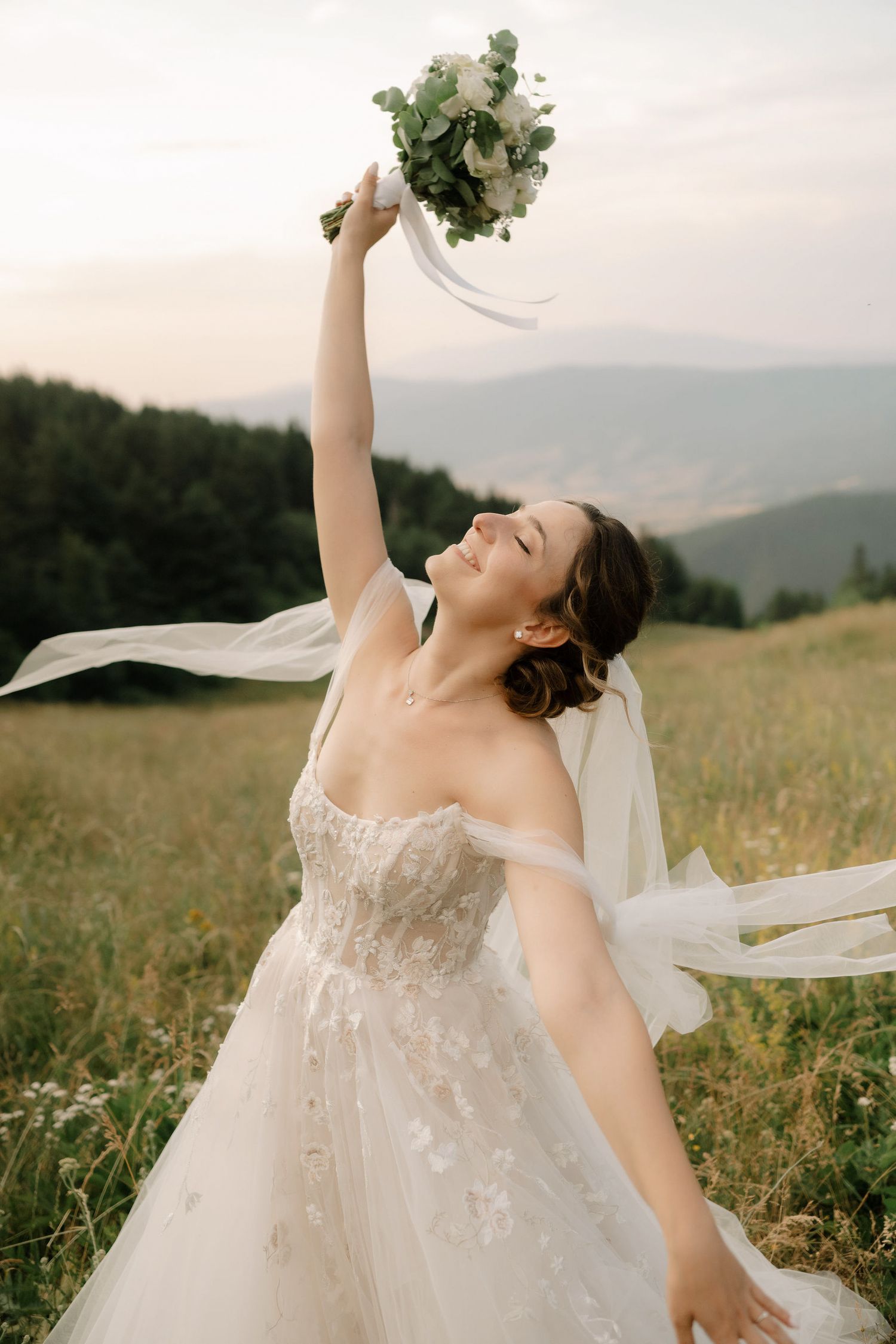 A joyful bride in a flowing white gown spins and dances in a sunlit meadow with mountains in the background.
