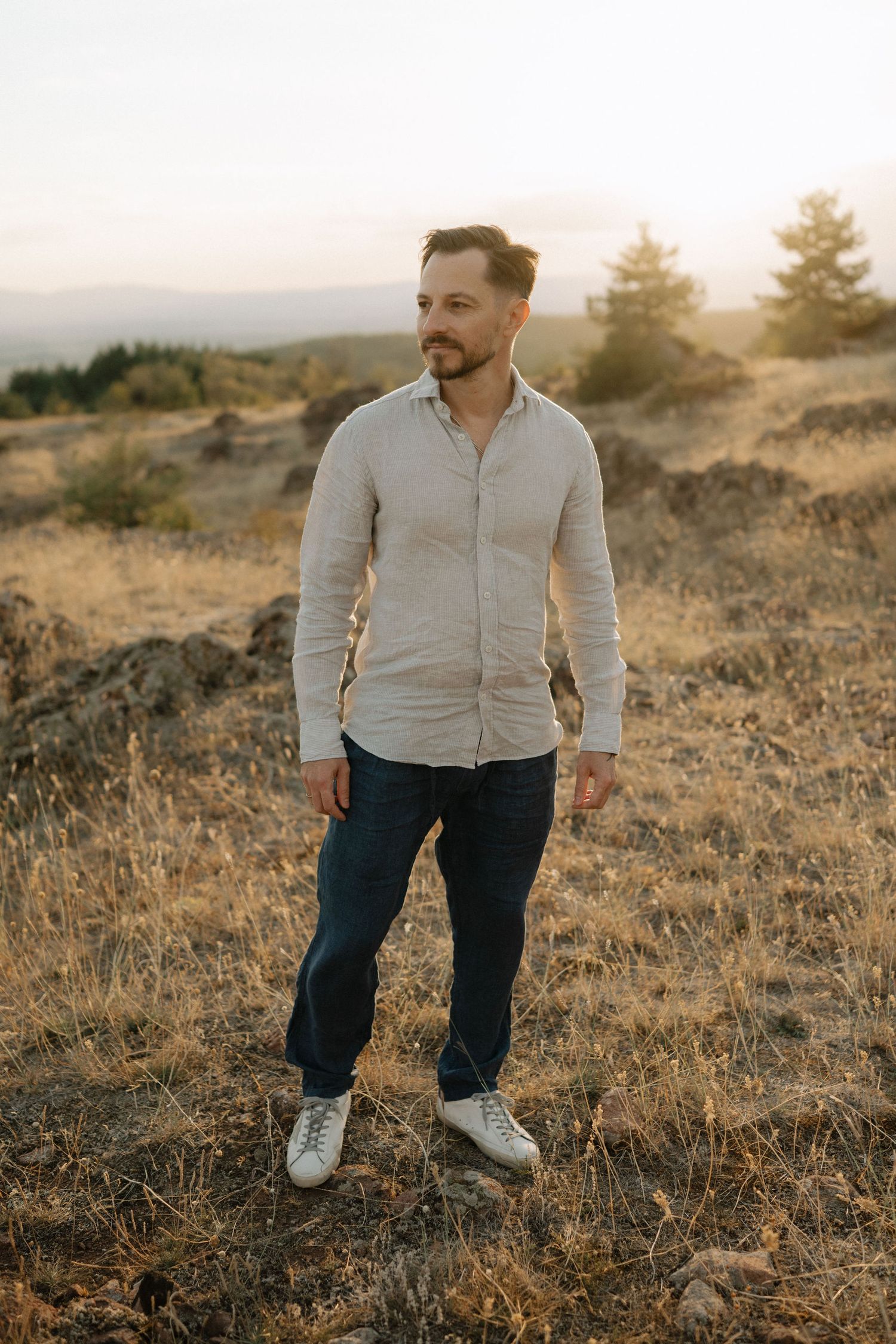 Person wearing a light gray zip-up jacket and dark jeans stands in a golden field at sunset.