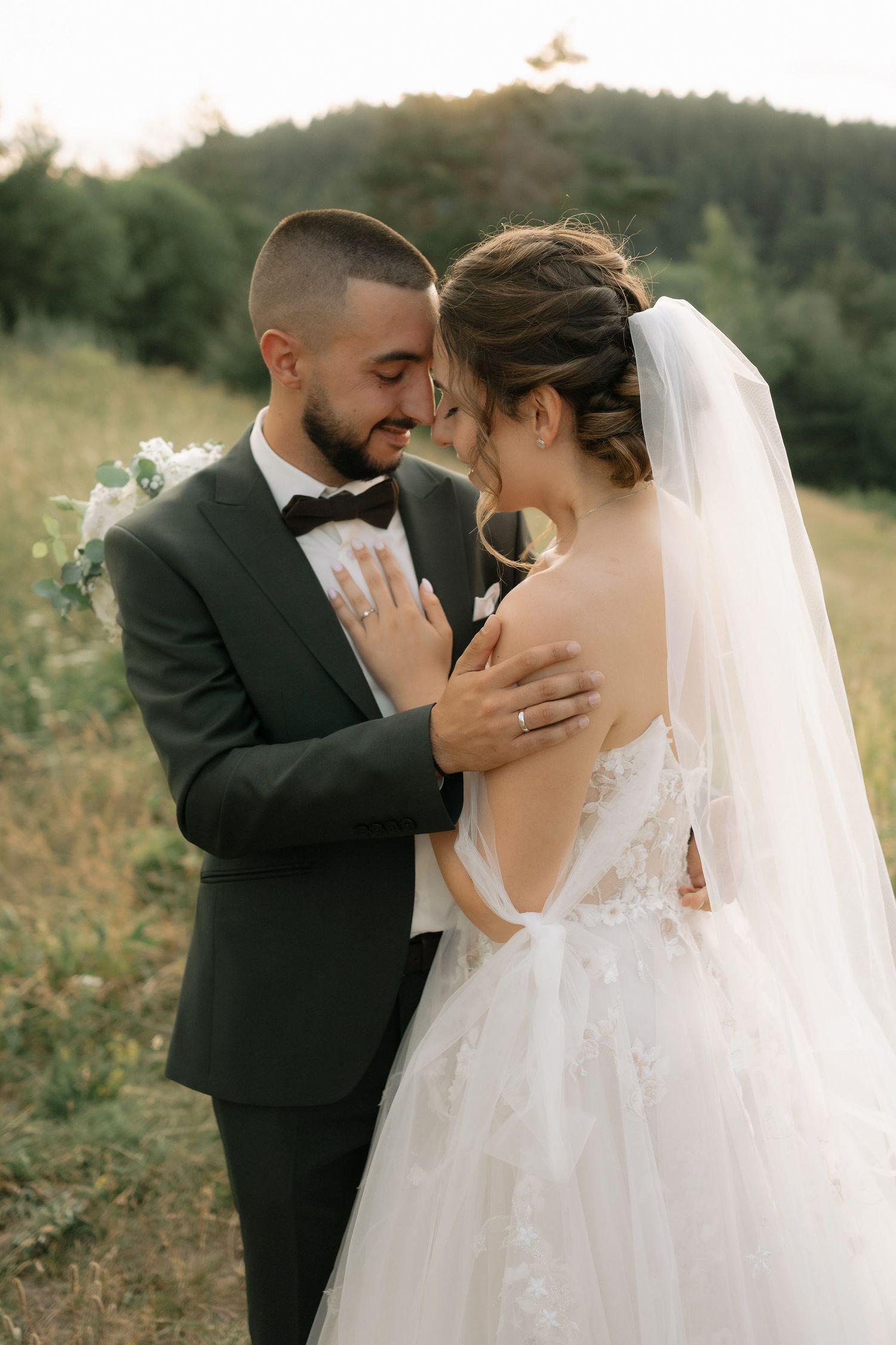 Romantic wedding couple embraces in a sunlit meadow with flowing white veil and black suit.