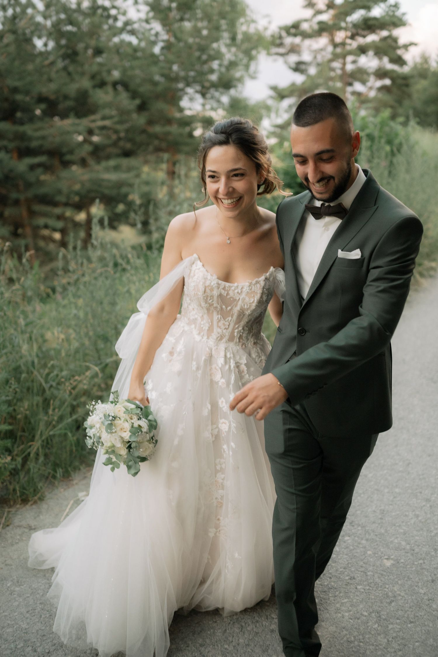A couple in wedding attire share a candid moment walking together outdoors on a path surrounded by greenery.