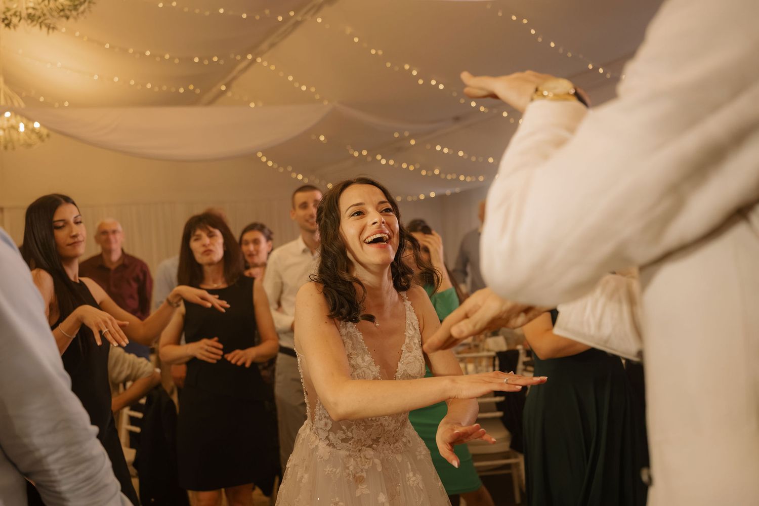 Bride laughing joyfully on the wedding dance floor surrounded by guests under warm fairy lights.
