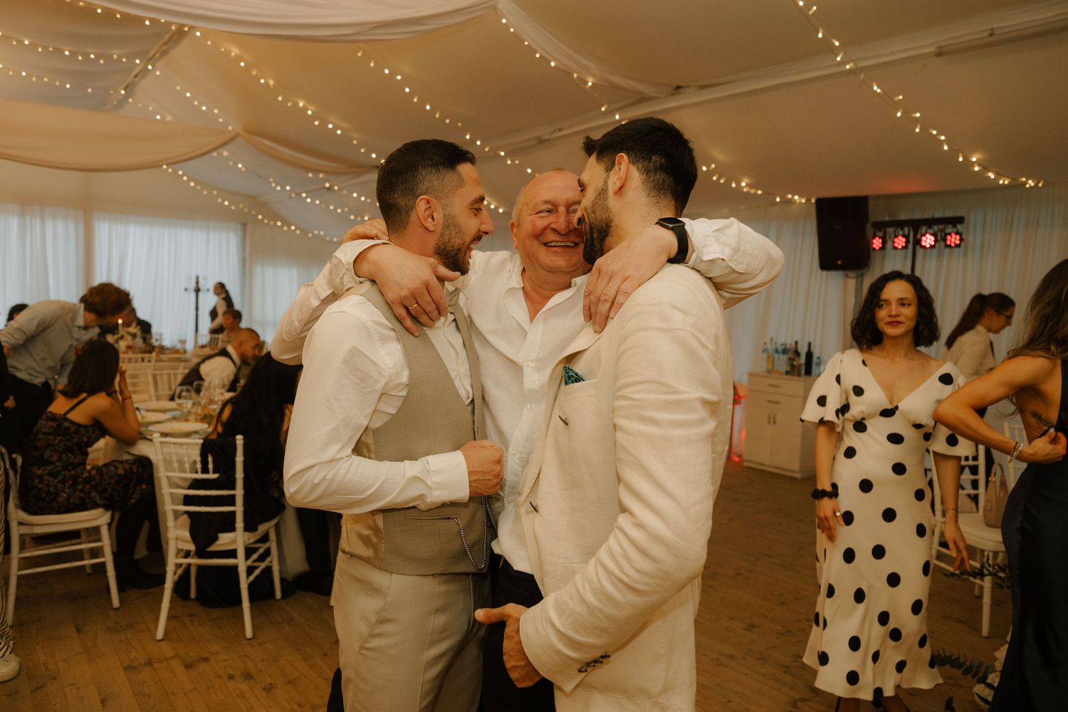 Three men share an emotional embrace on a wedding dance floor decorated with fairy lights.