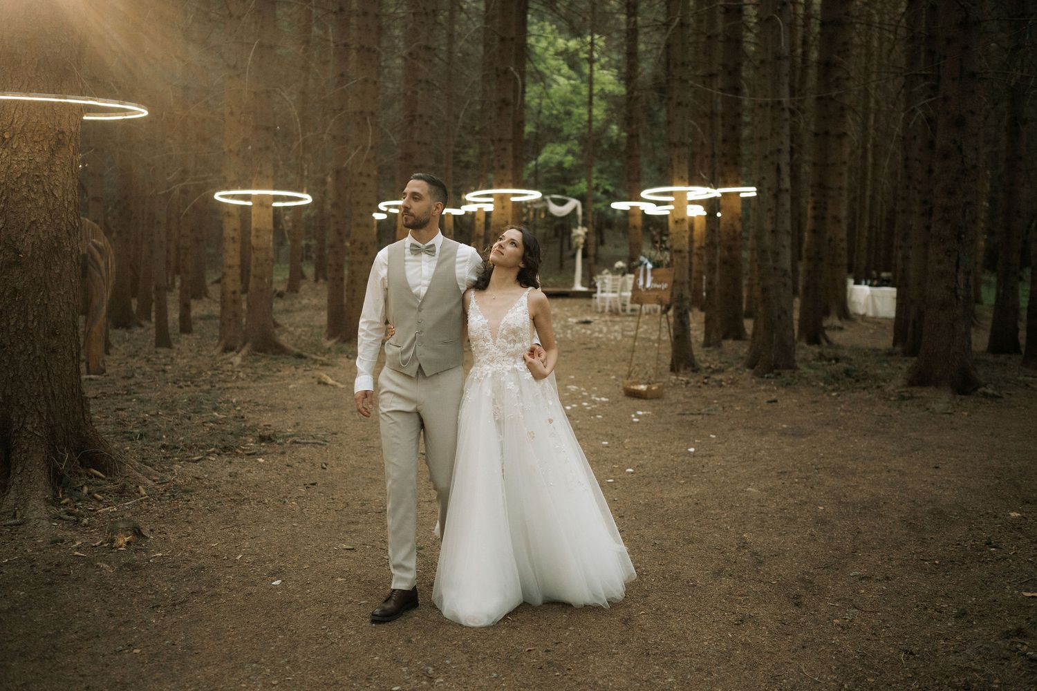 Bride and groom walk hand in hand through a forest wedding venue adorned with glowing circular chandeliers.