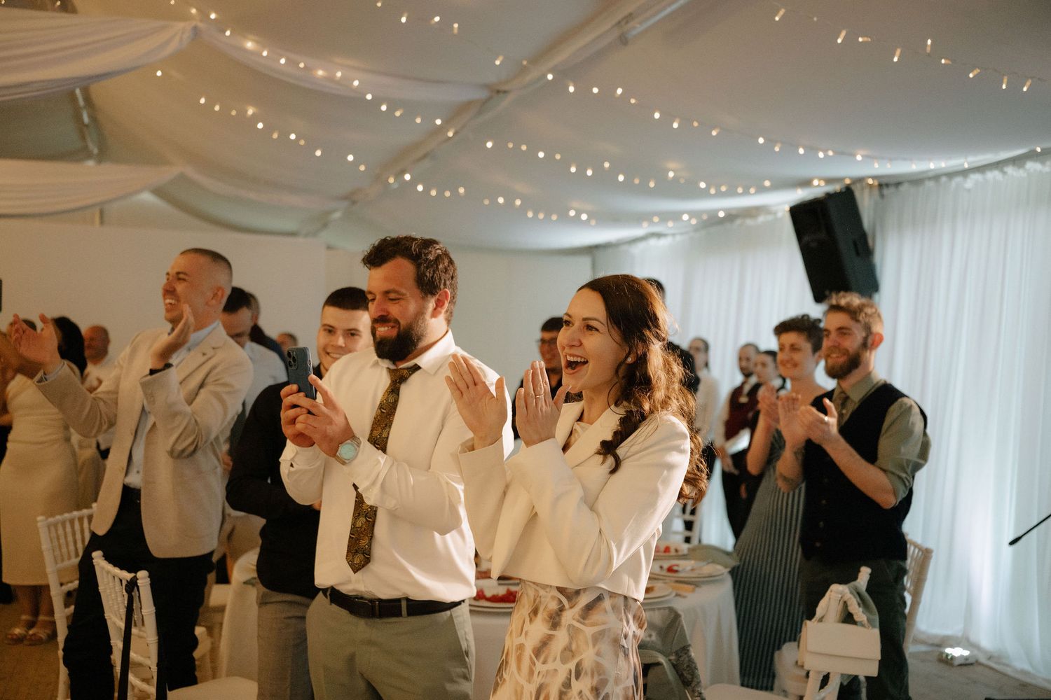 Guests clapping and laughing at a wedding reception inside a decorated marquee with fairy lights.