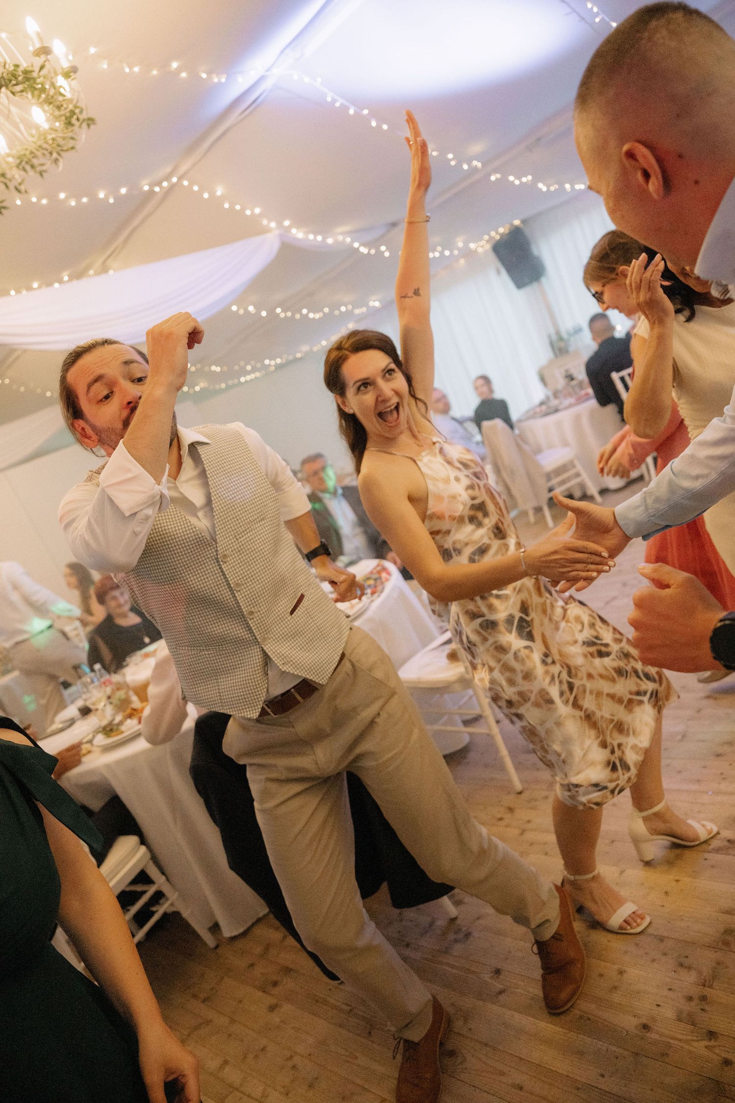 Guests dancing joyfully at a wedding reception in a decorated marquee with fairy lights and white draping.