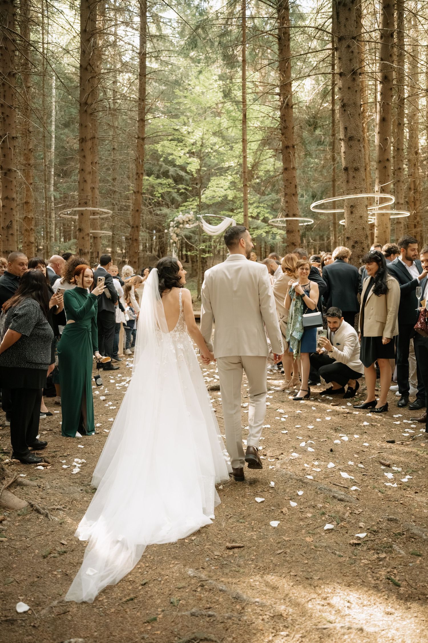 Bride and groom walk hand-in-hand down a forest aisle surrounded by guests during an outdoor woodland wedding ceremony.