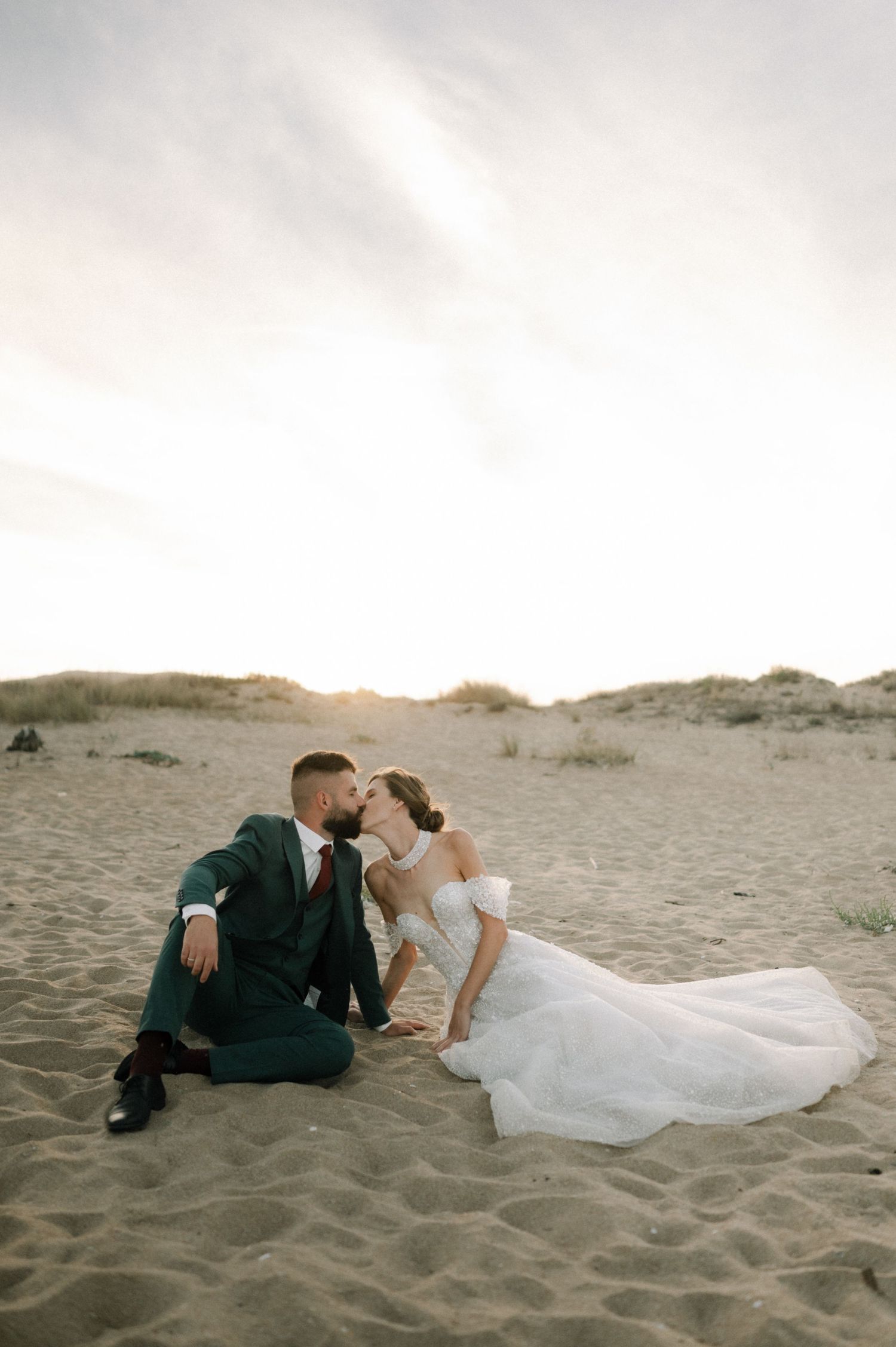 A bride and groom share a romantic kiss while sitting on sandy beach dunes at golden hour sunset.