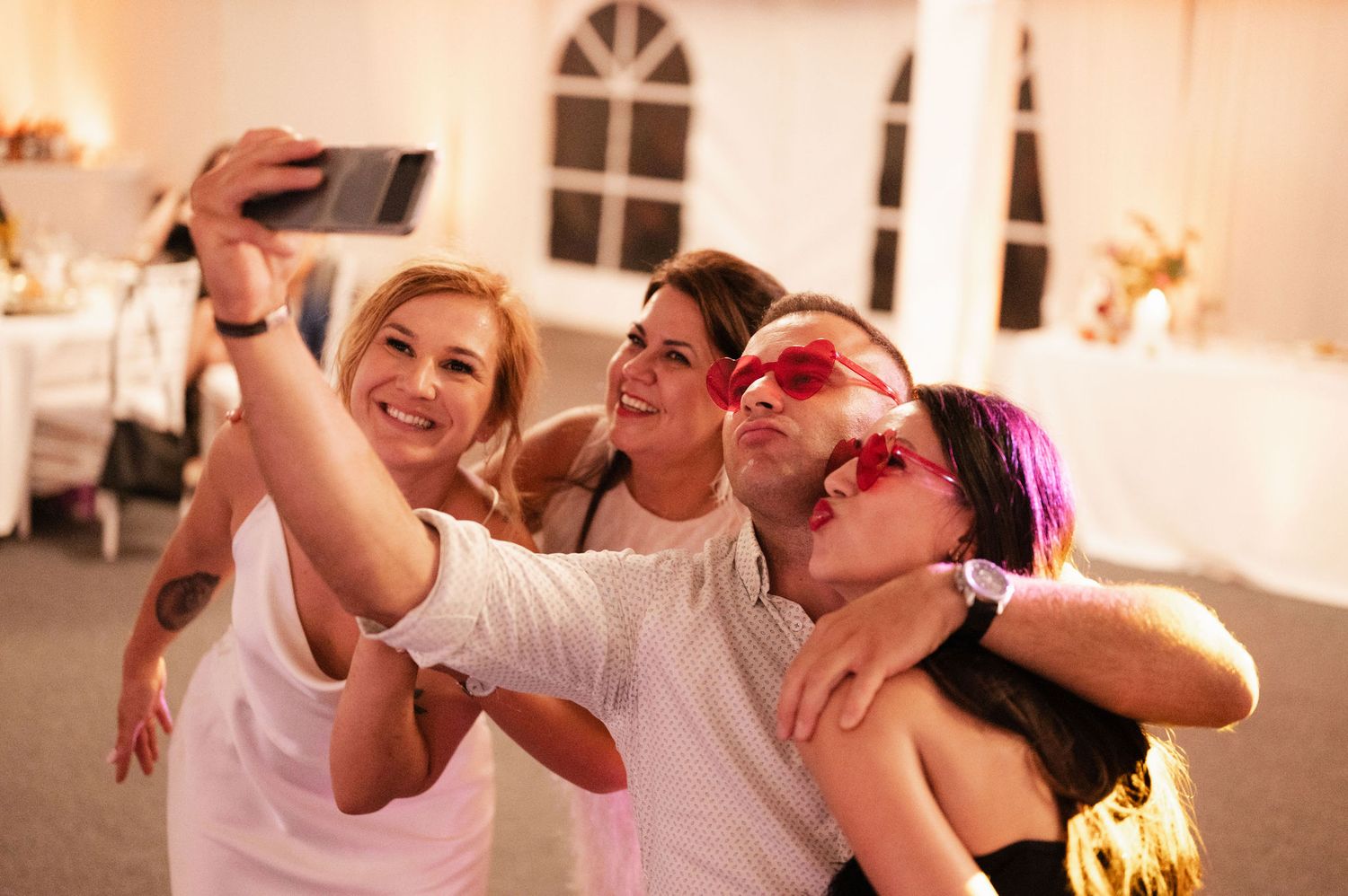 Group of friends taking selfies and making silly faces at a party with warm lighting in the background.
