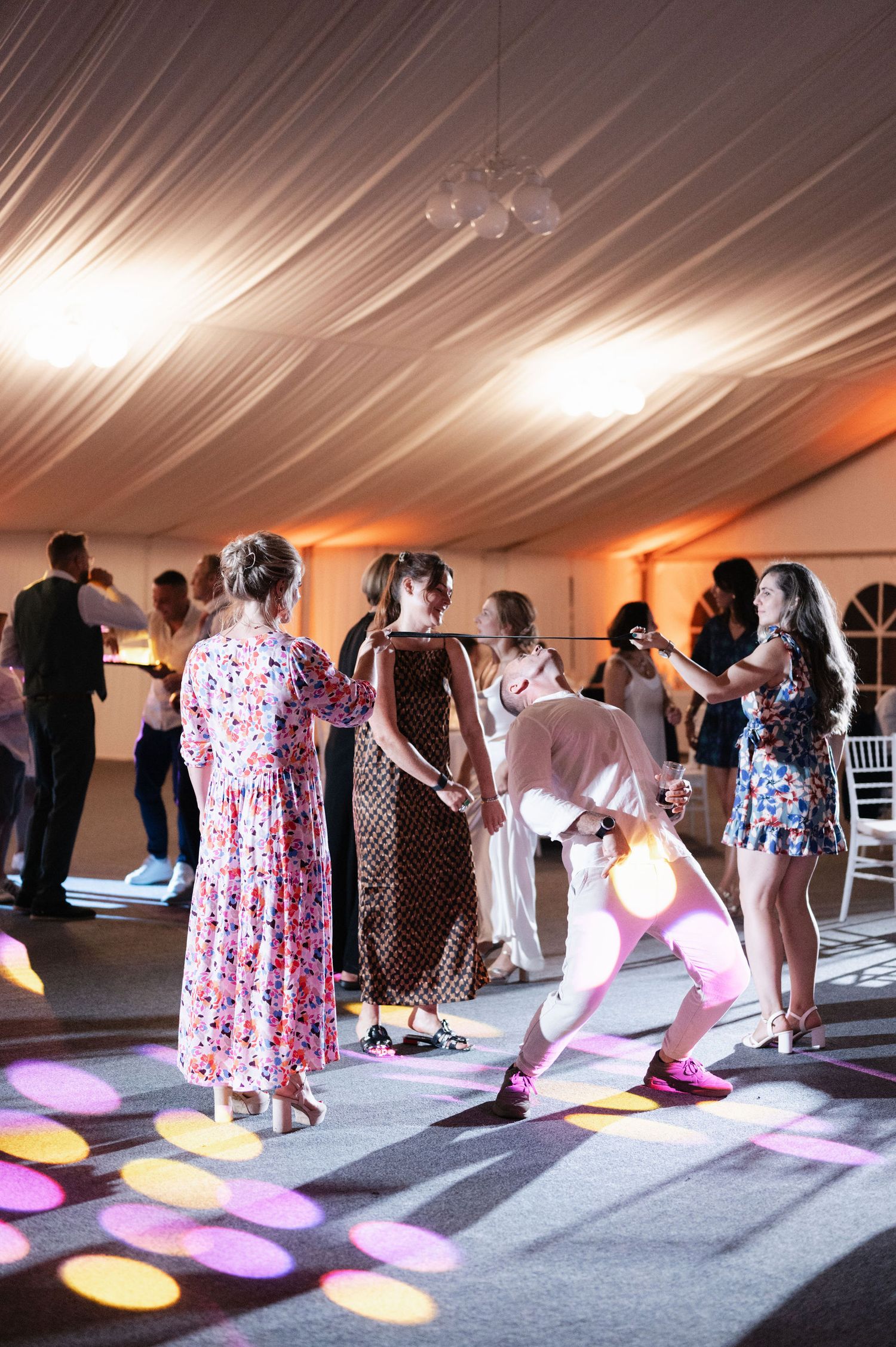 People dancing in a tent venue with colorful spotlights casting pink and yellow circles on the floor.