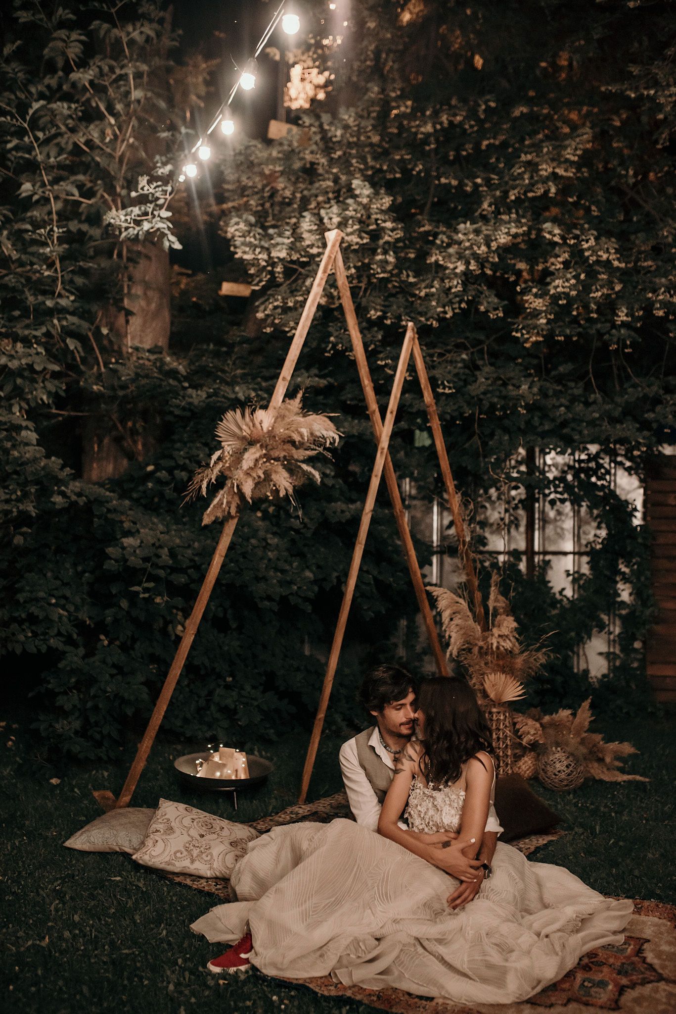 A romantic outdoor evening scene with glowing lights and a couple reclining on blankets in a wooded setting.