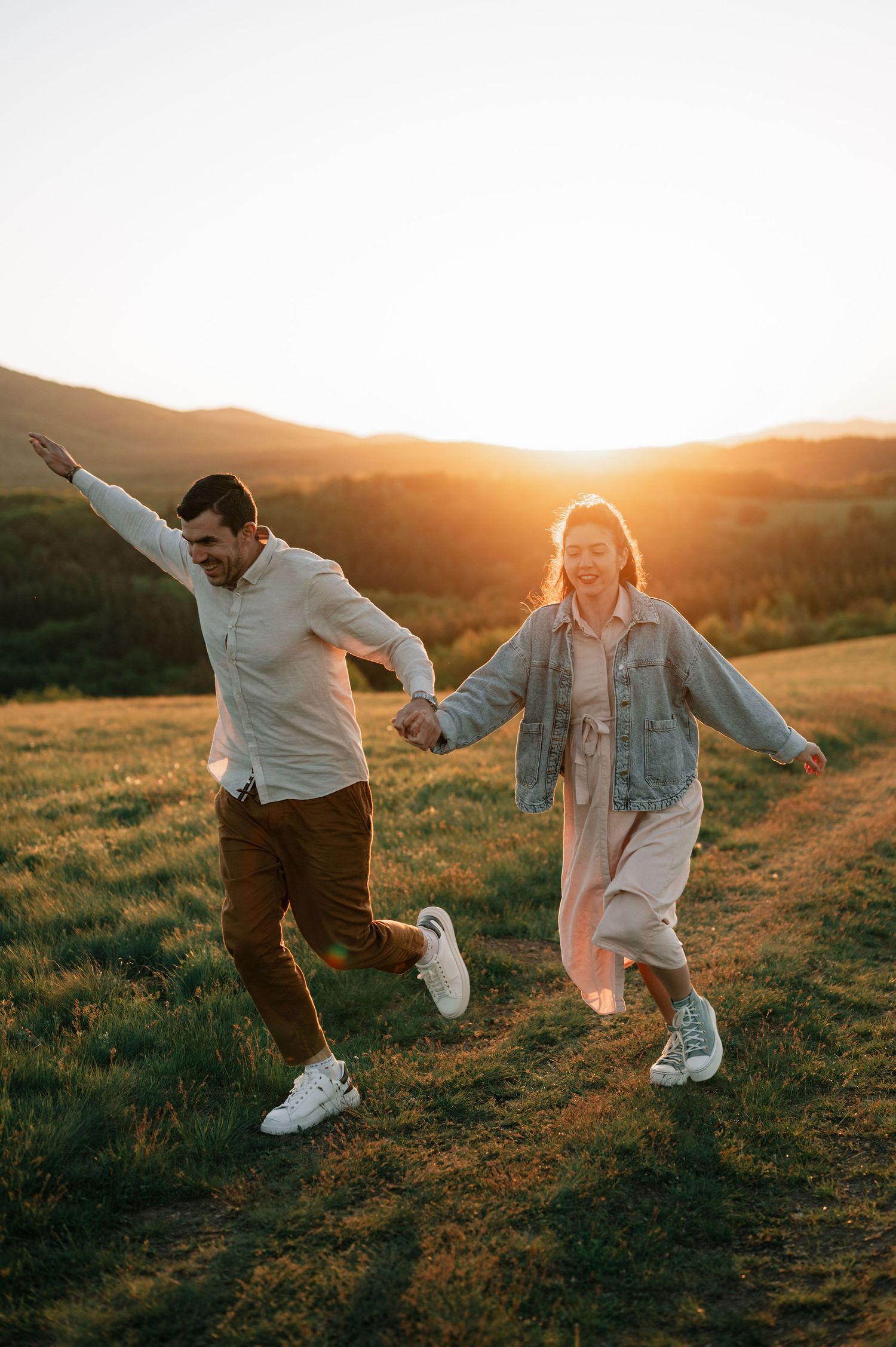 A couple playfully runs and dances together on a hilltop during golden hour sunset.